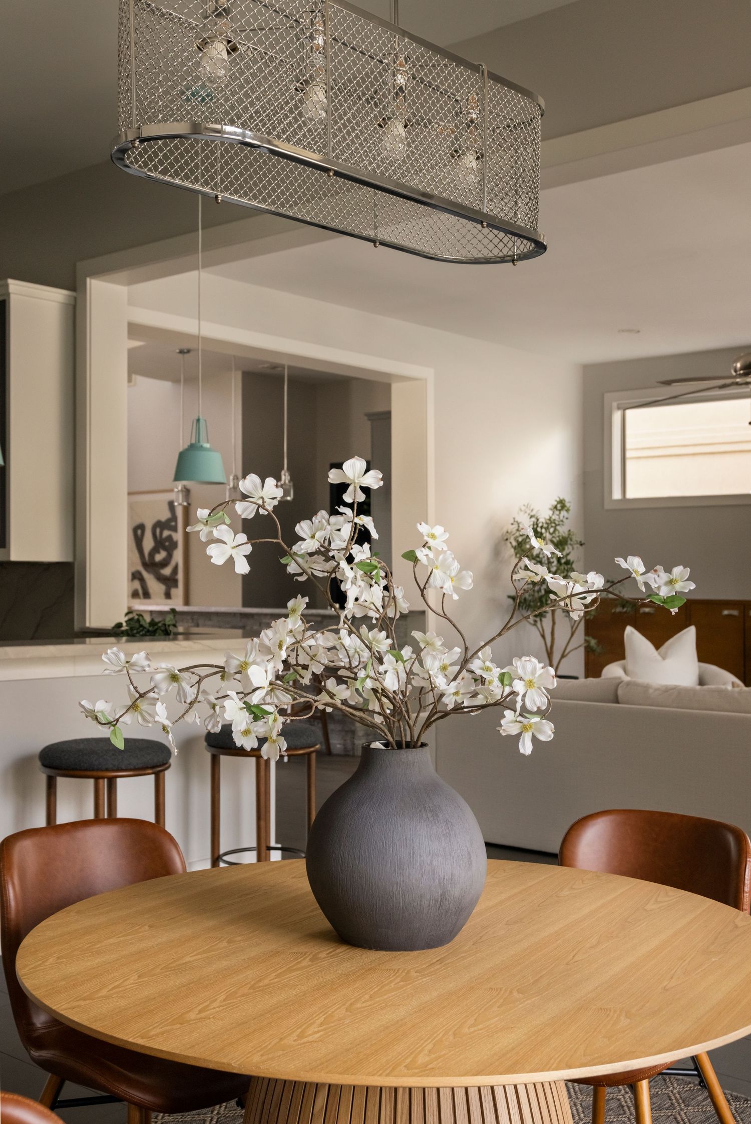 Contemporary dining space with round oak table, mesh pendant light, and gray vase with white flowers.