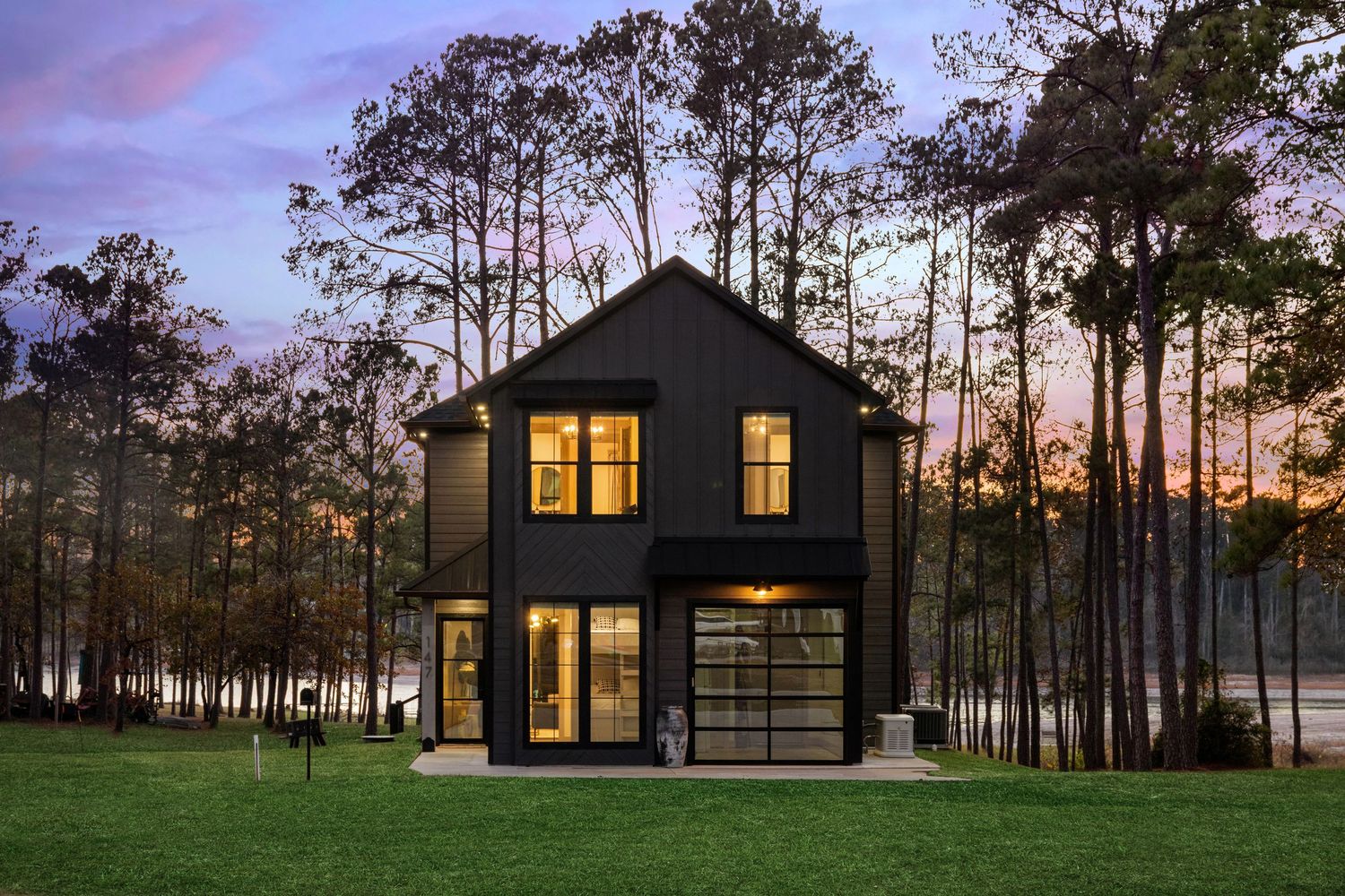 Modern black house with large windows illuminated at dusk among tall pine trees.