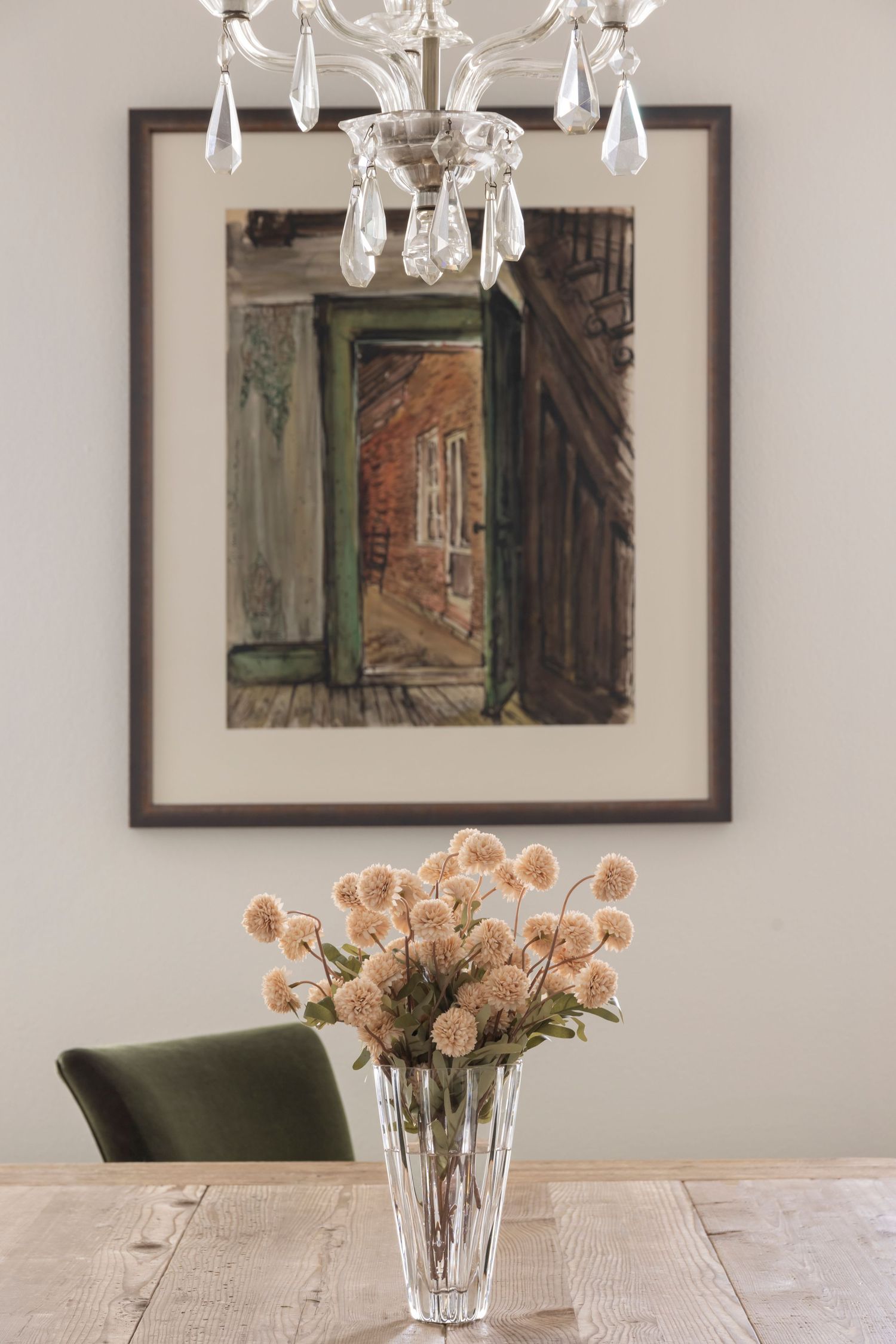 Crystal chandelier hangs above rustic wooden table with peach roses in vase and framed artwork on white walls.