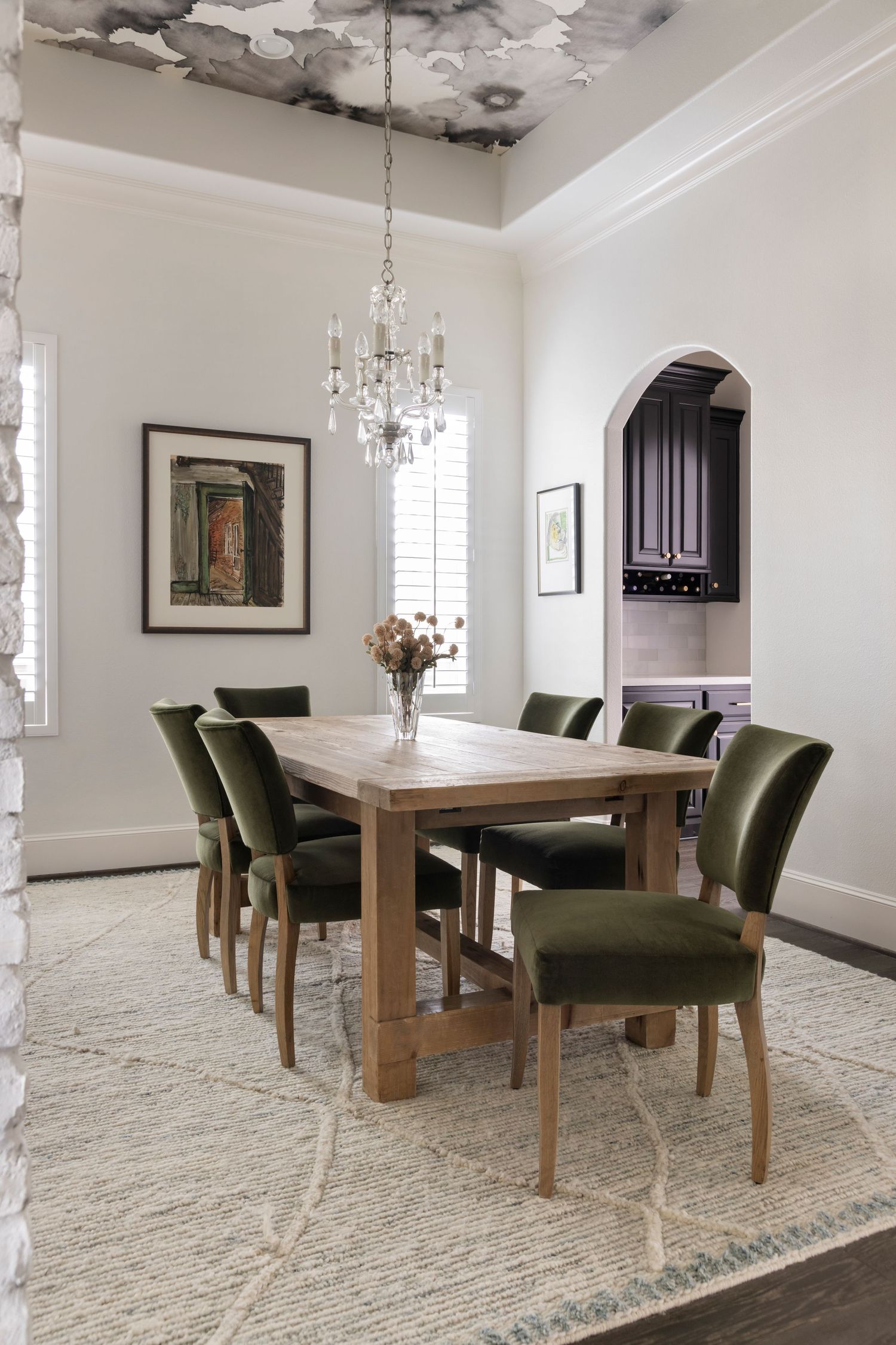 Modern dining room with wooden table, olive green chairs, crystal chandelier and cream carpet on white backdrop.