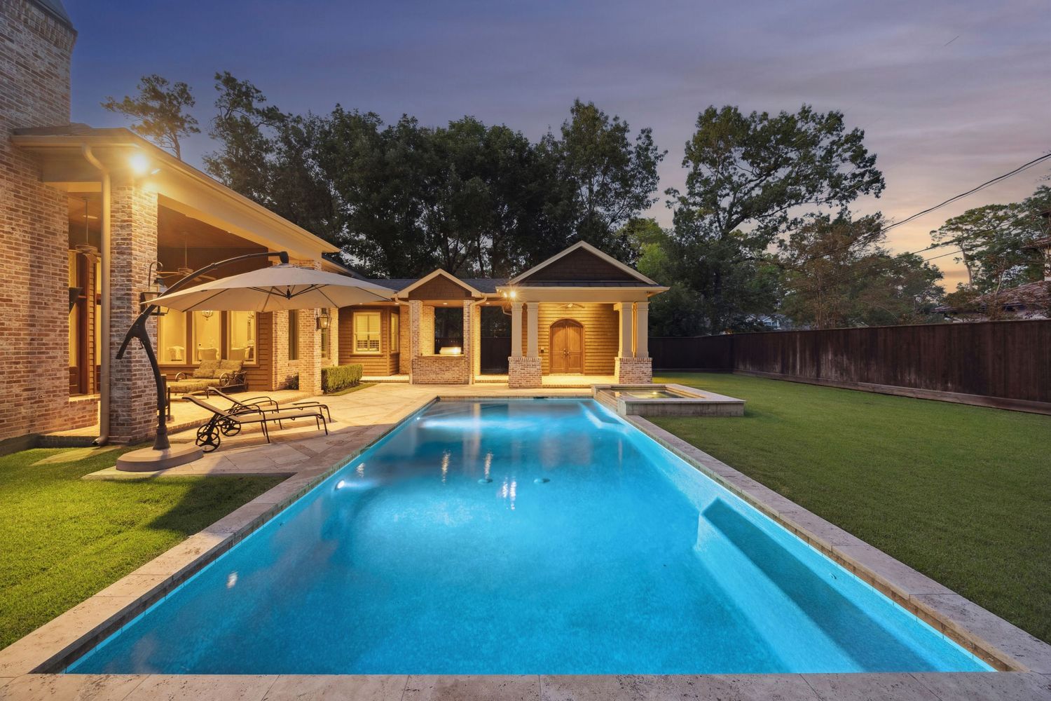 Modern luxury home exterior with illuminated swimming pool and landscaped yard at sunset.