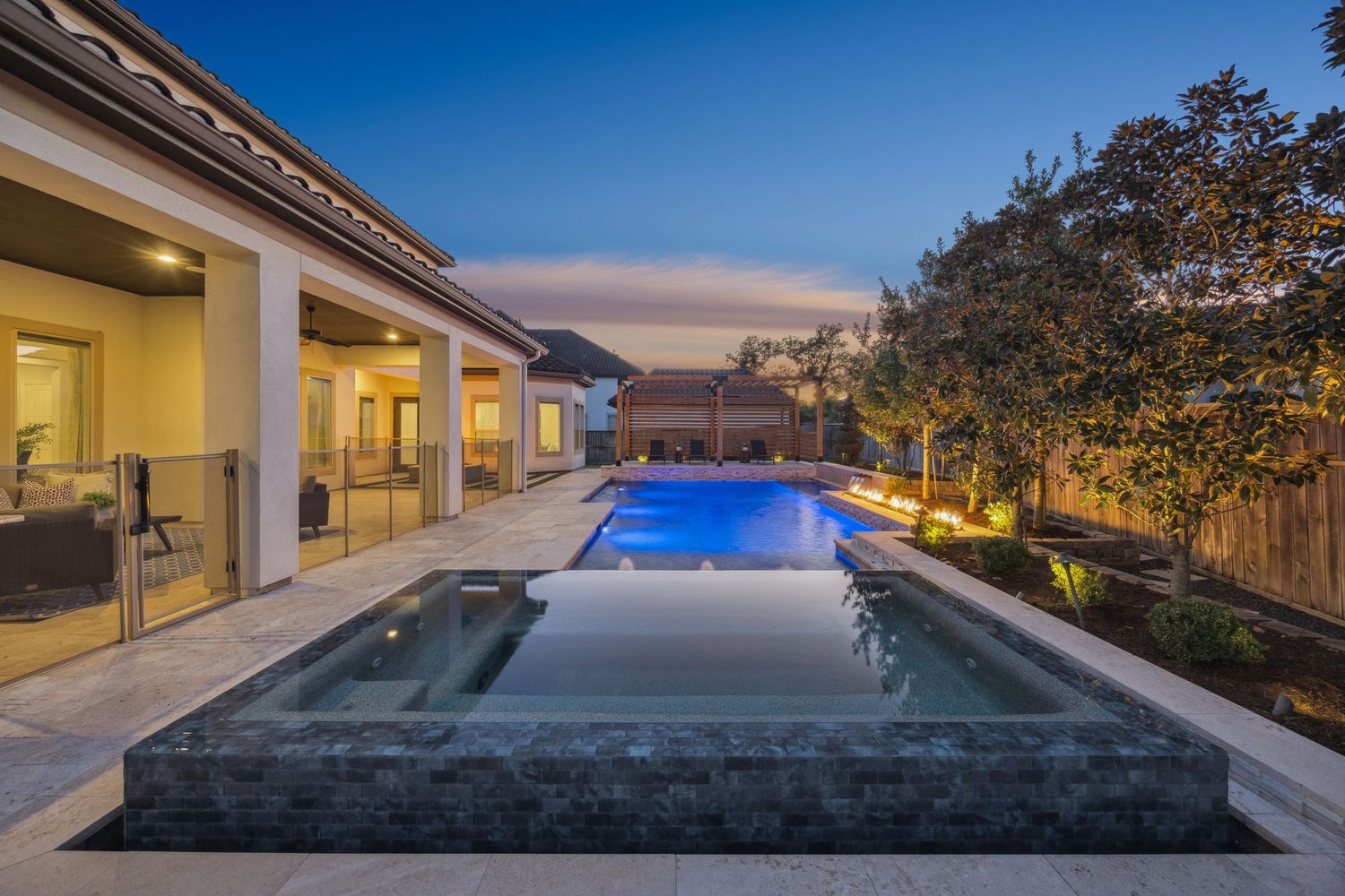 Luxury home exterior features illuminated infinity pool and covered patio at sunset.