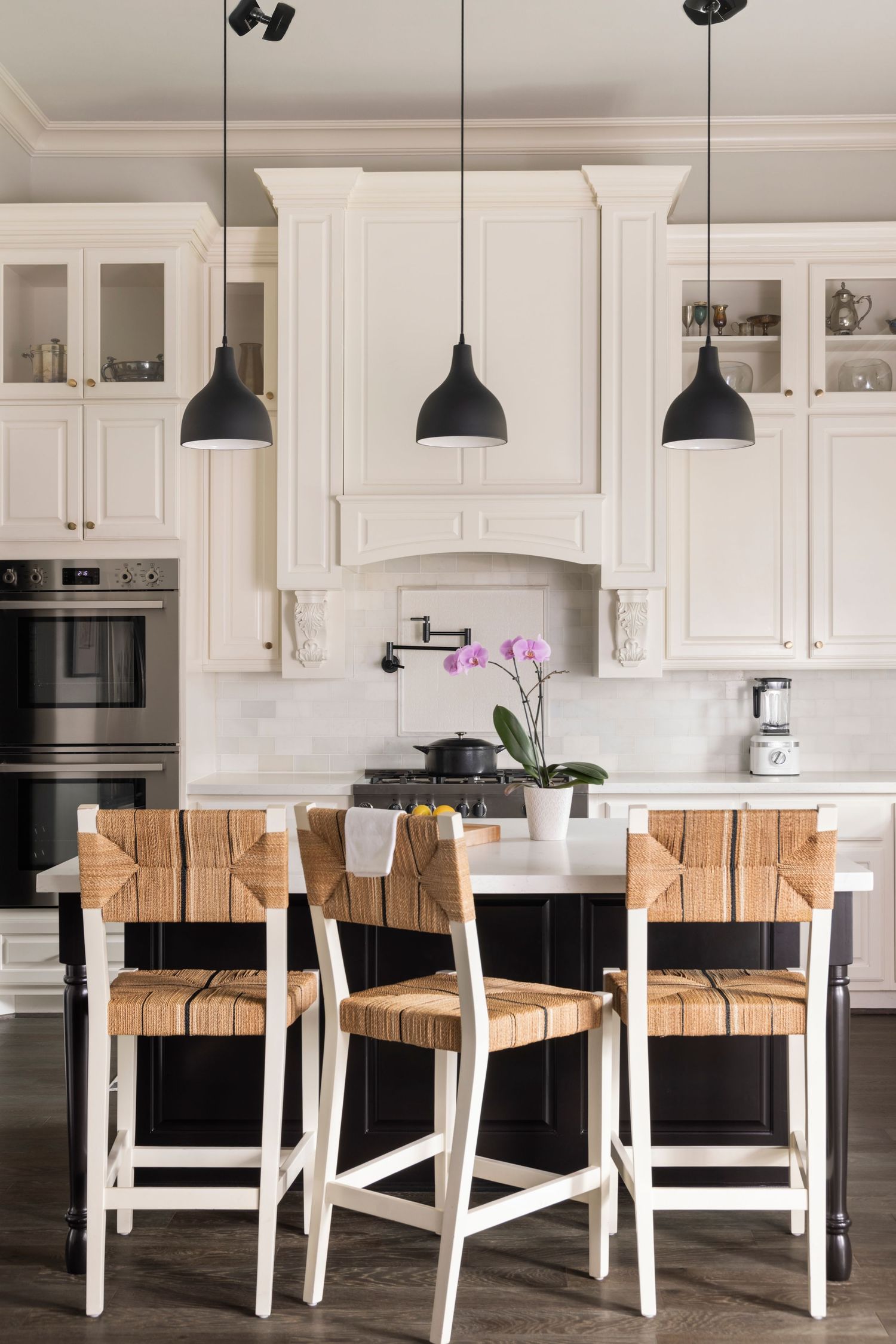 Modern white kitchen with black pendant lights and woven leather counter stools featuring white and natural wood tones.