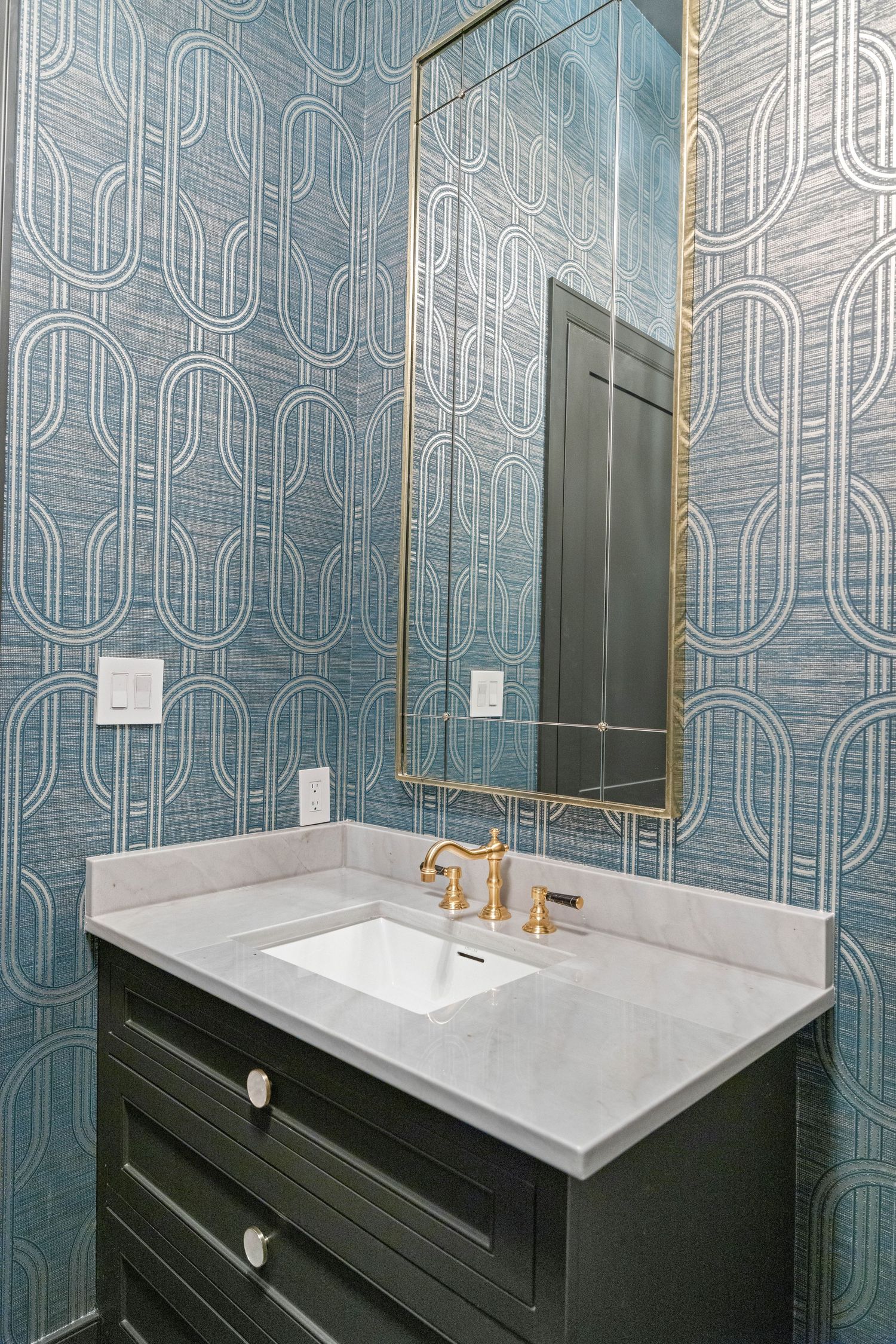 Modern bathroom featuring geometric blue wallpaper, black vanity, and gold-framed mirror.