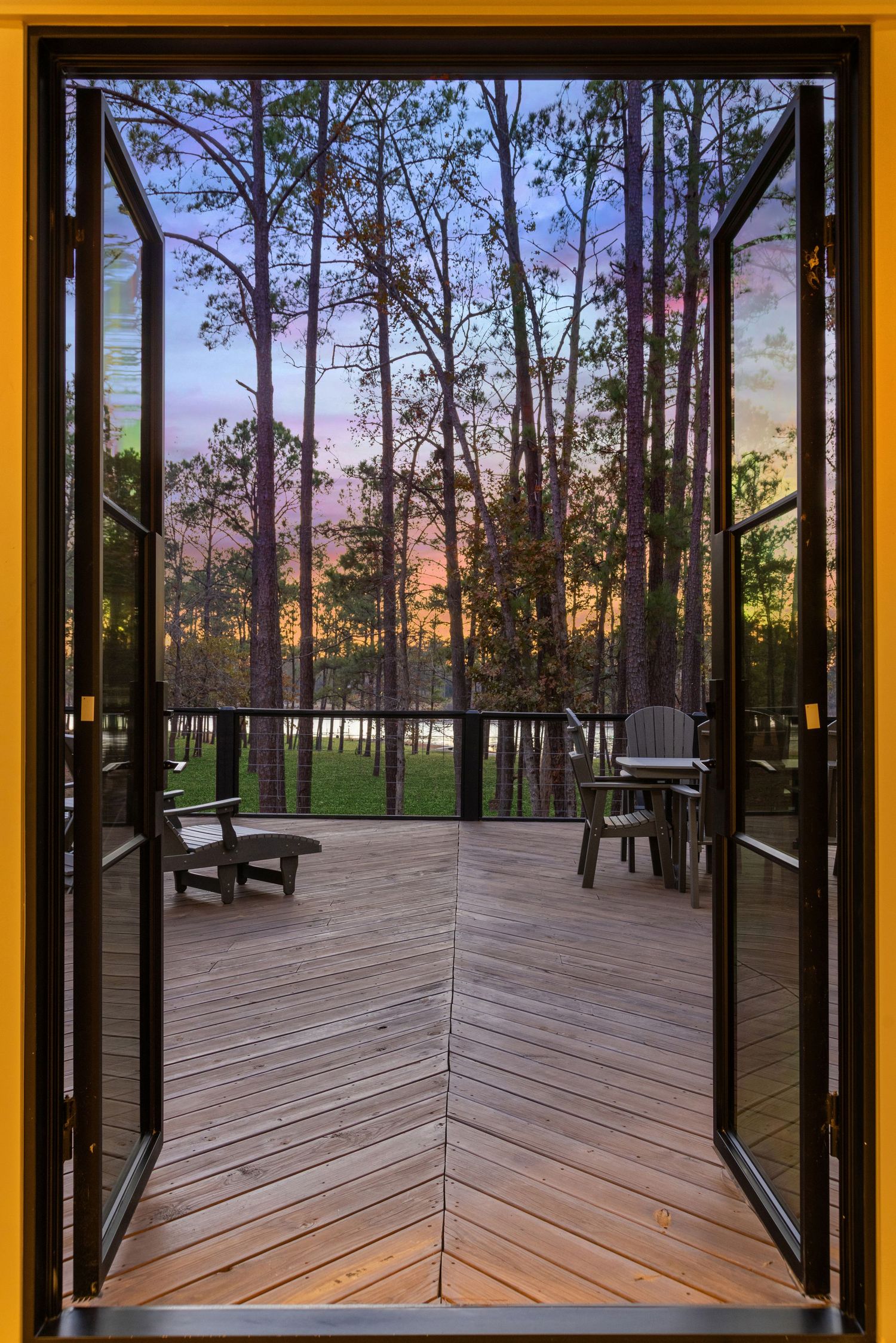 Glass doors frame a symmetrical view of wooden deck and pine forest at dusk.