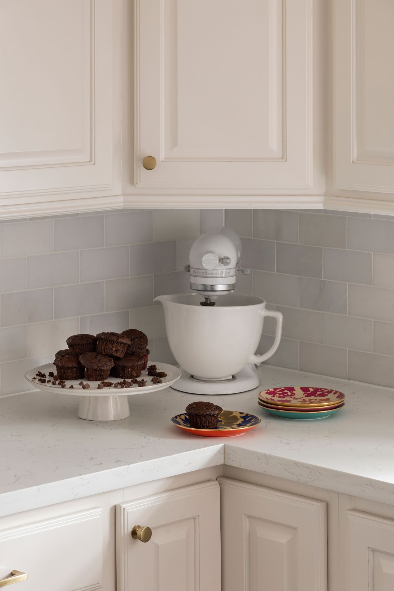 White KitchenAid stand mixer in corner of kitchen counter with chocolate cookies on cake stand and colorful plates.