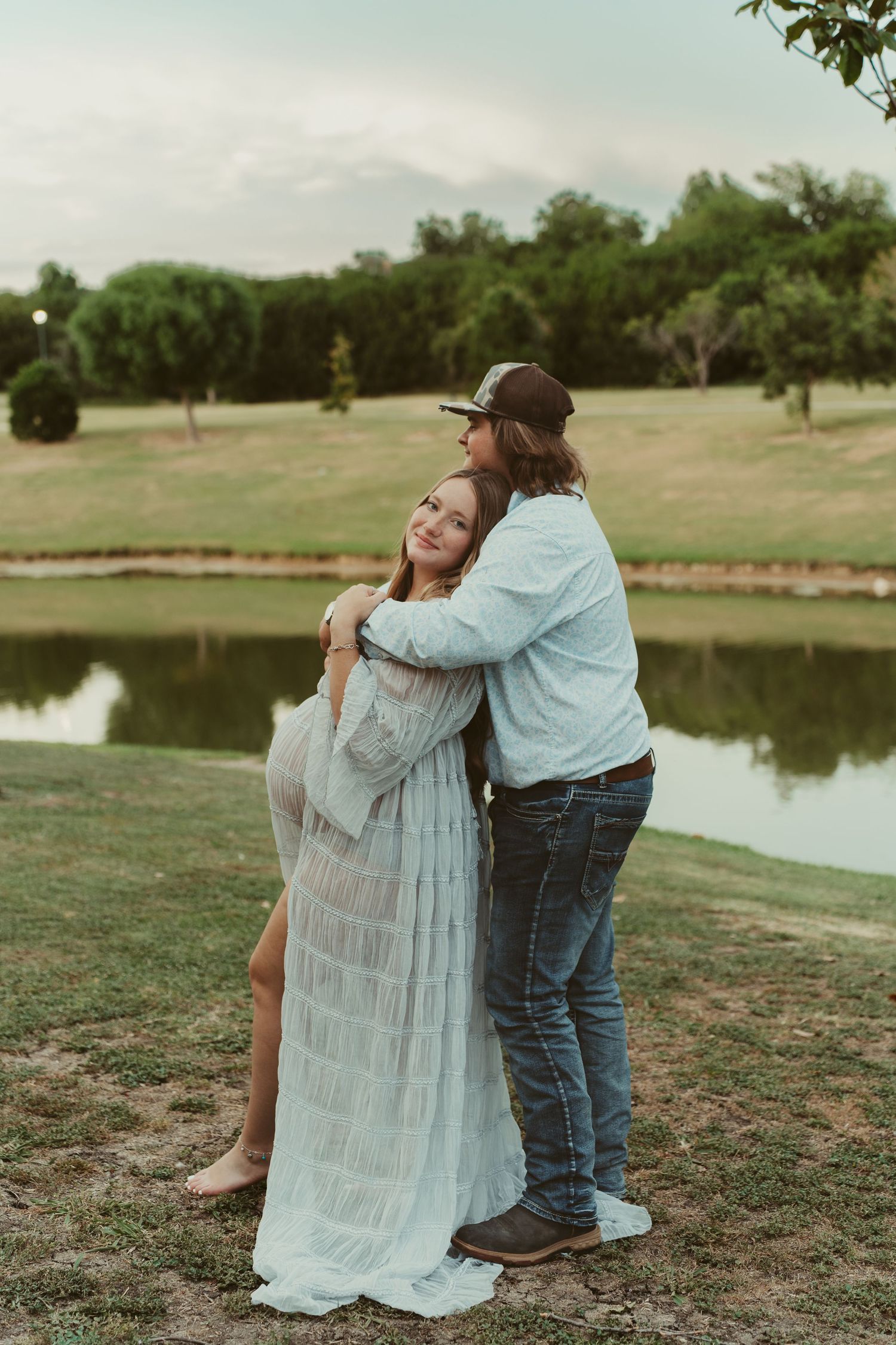 A couple embraces outdoors near a pond during a maternity photo session.