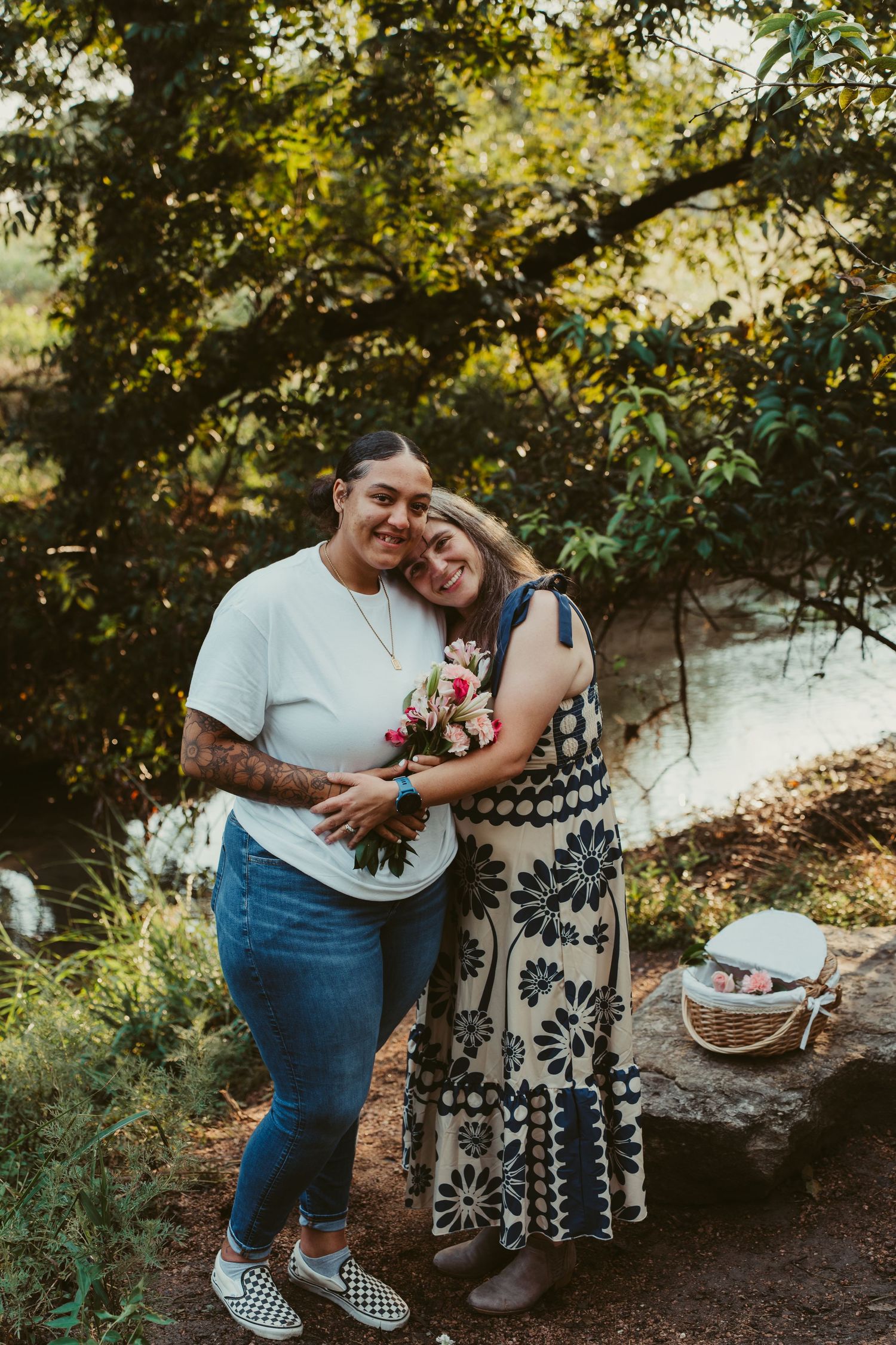 Two women embrace and smile while holding a bouquet of flowers during an outdoor celebration.
