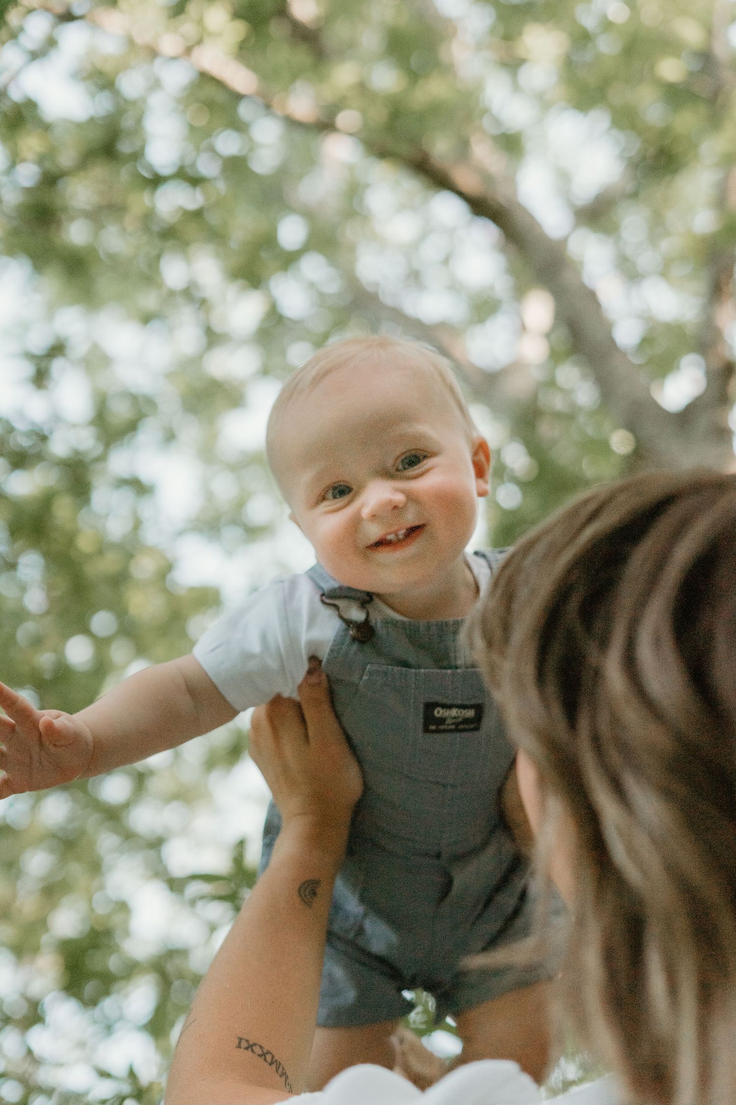 Smiling baby in denim overalls lifted up by mother beneath leafy green trees.