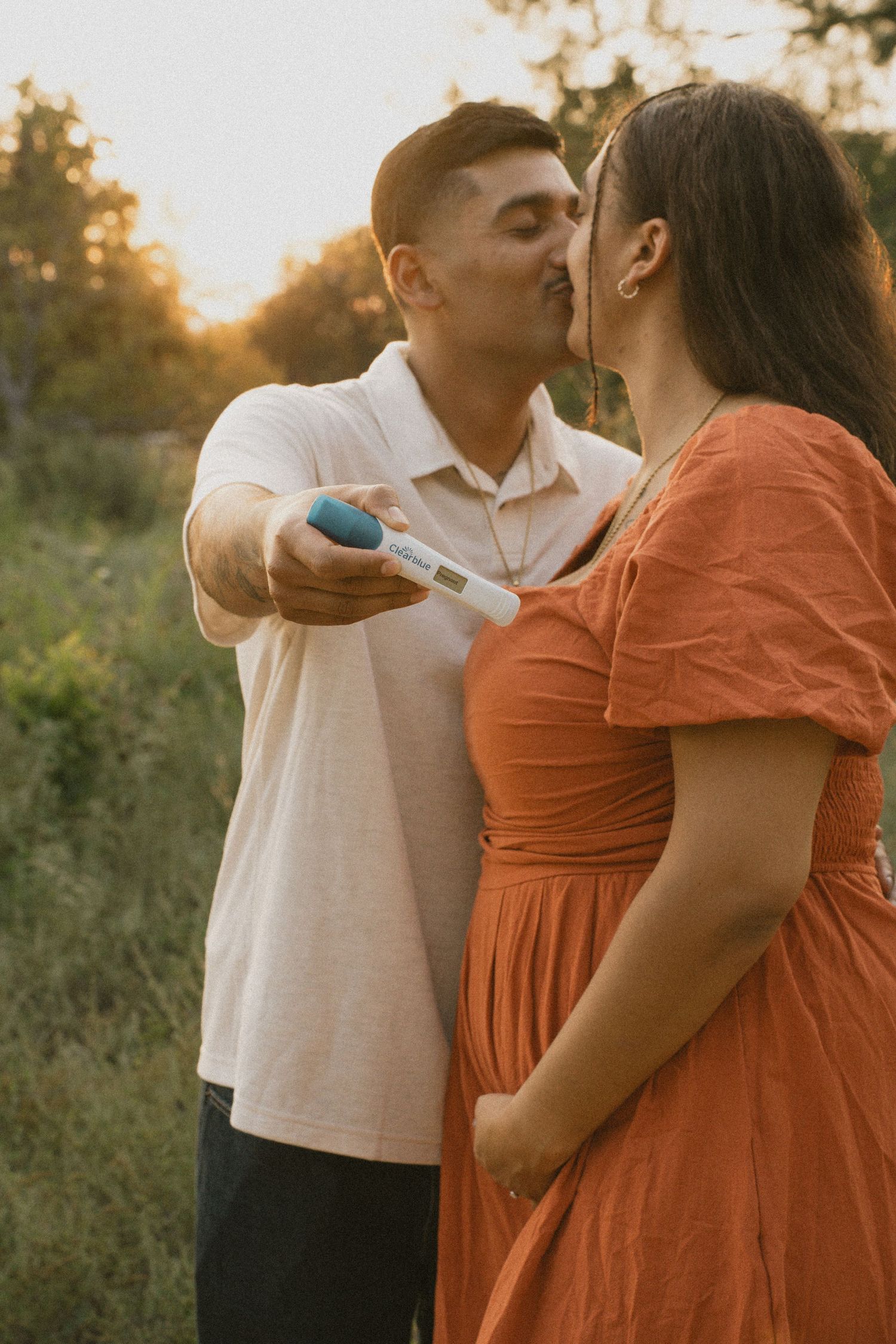 Couple kissing at sunset while holding a positive pregnancy test to announce pregnancy.