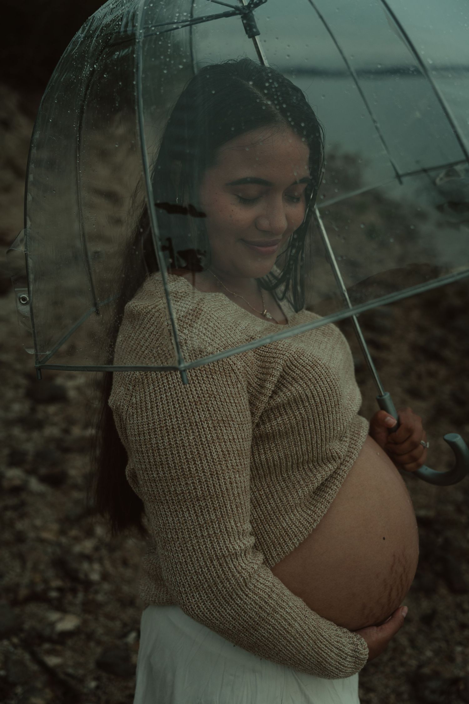 Smiling pregnant woman sheltering under transparent umbrella in rain, gently holding her belly.