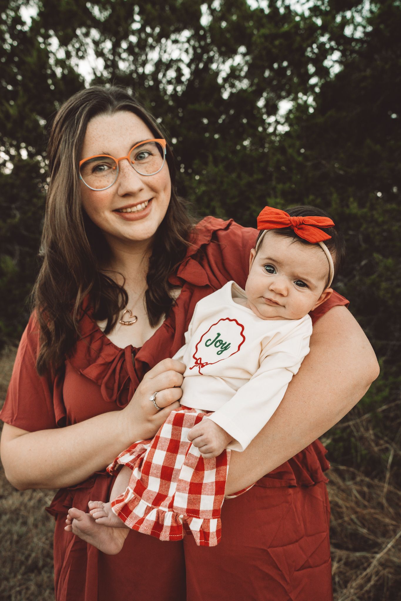 Smiling mother with orange glasses holding baby girl in red gingham dress outdoors.