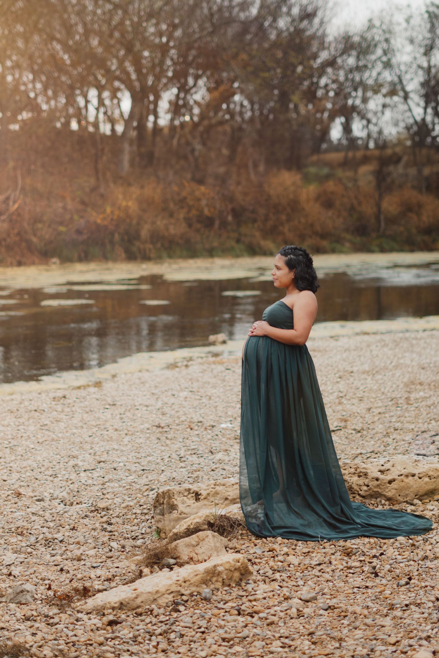 Elegant pregnant woman in flowing teal gown stands by rocky riverbank during maternity photo session.