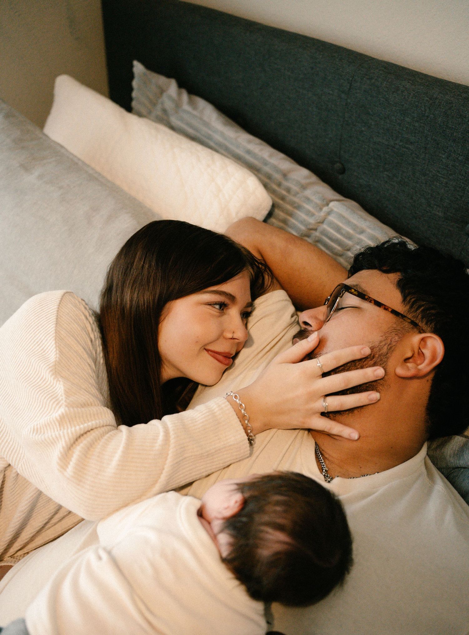 Loving couple smiling at each other on bed with newborn baby in warm home setting.