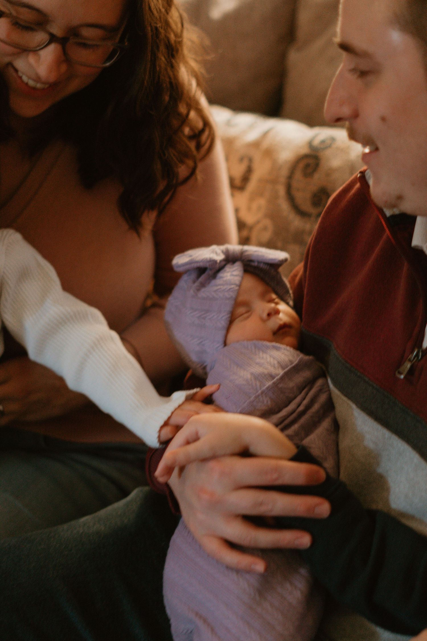 Smiling parents holding sleeping newborn wrapped in purple swaddle on couch.