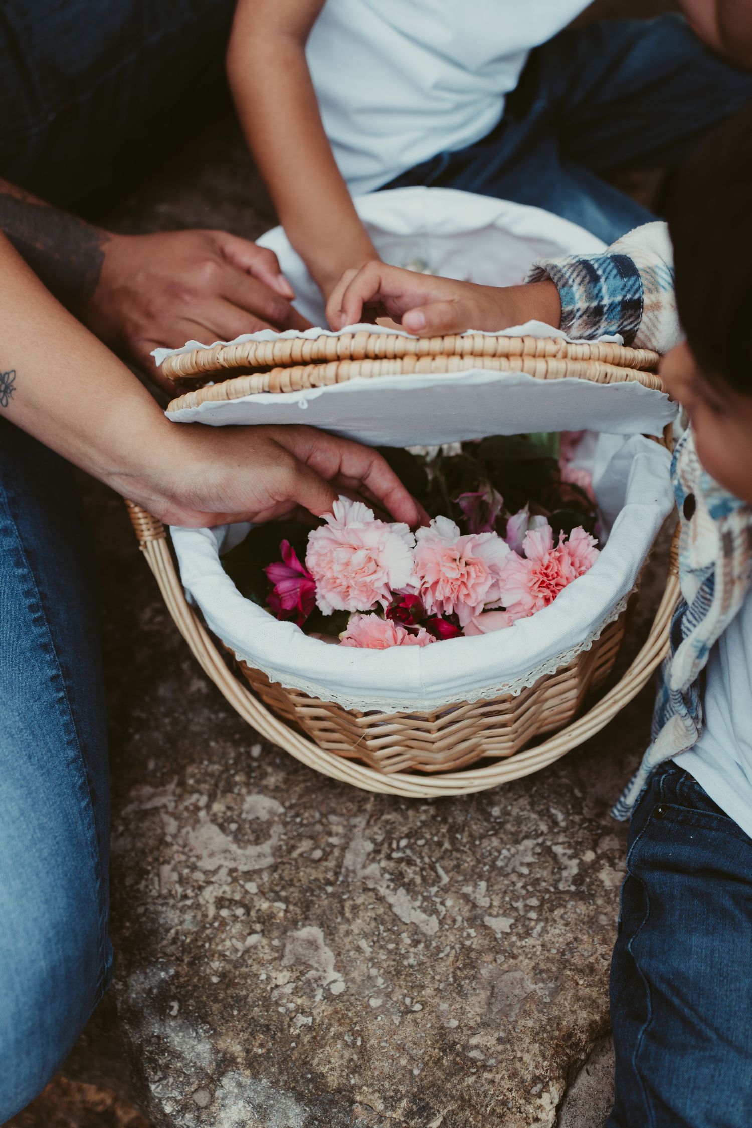 Close-up of hands opening a wicker basket filled with pink peonies and flowers outdoors.
