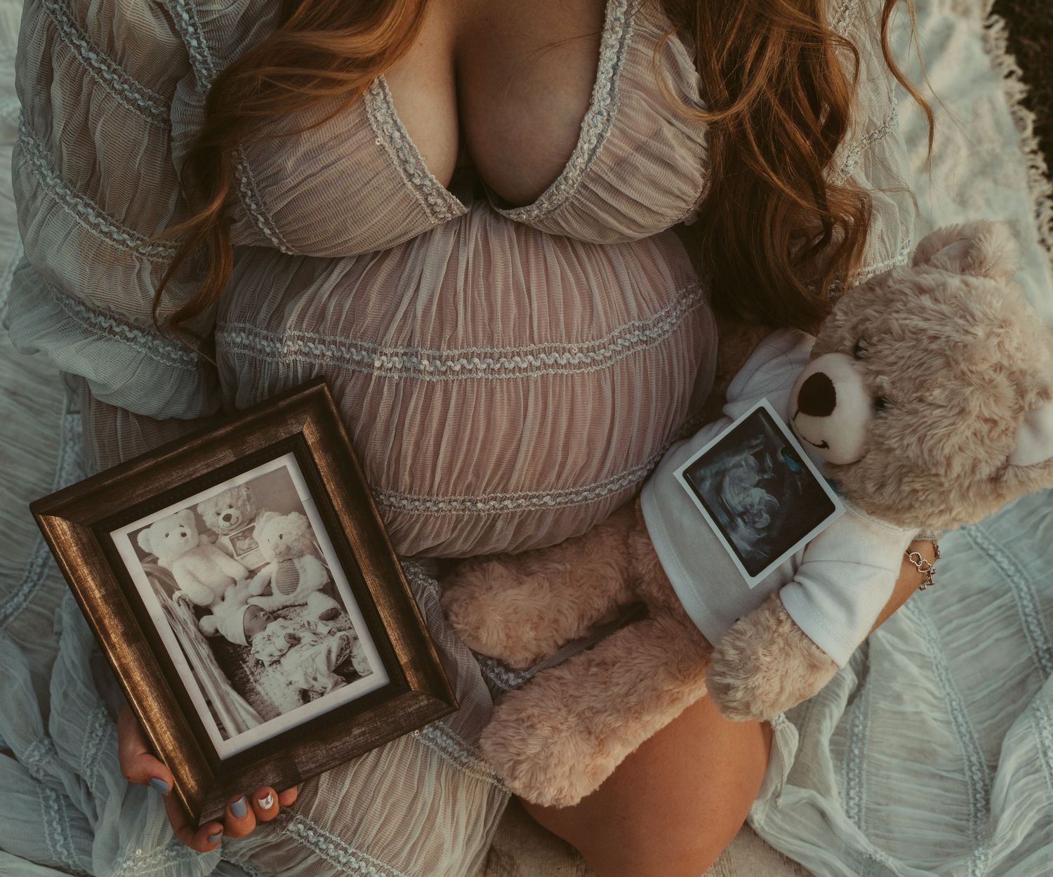 Pregnant woman in lace dress holding teddy bear and ultrasound photo while sitting on bed.