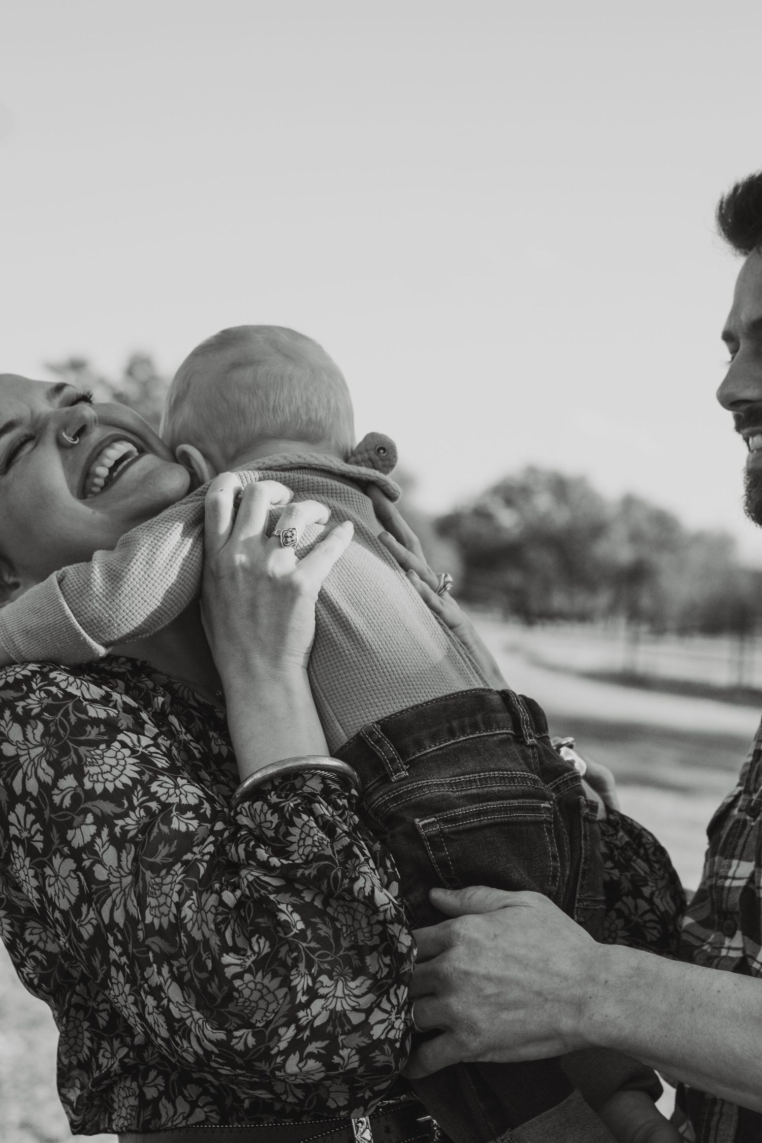 Black and white photo of joyful parents laughing while holding their baby outdoors.