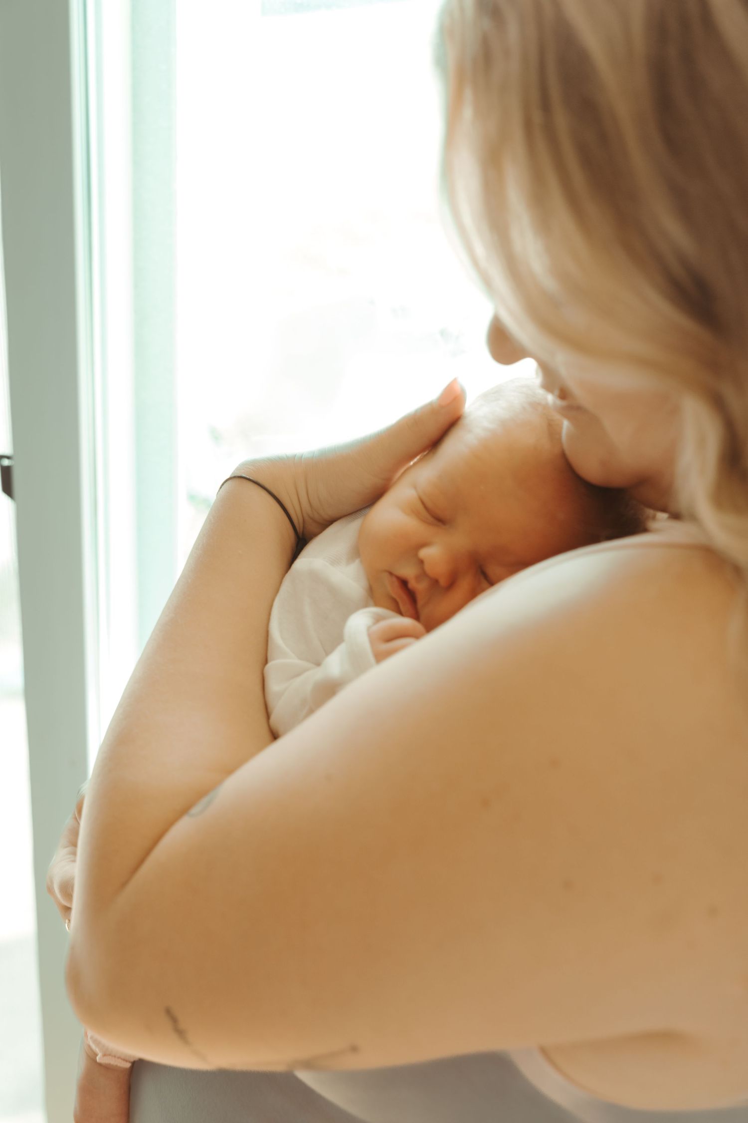 A blonde mother gently kisses her sleeping newborn baby while standing near a bright window.