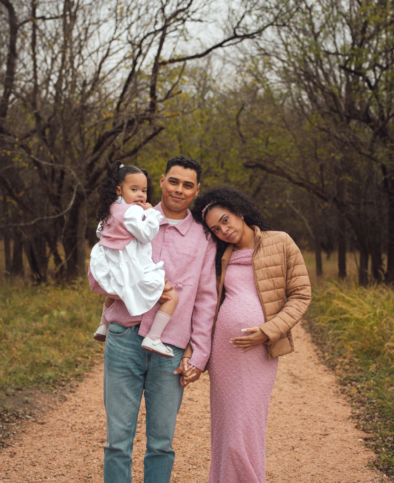 A maternity family portrait of a couple and toddler on a tree-lined dirt path outdoors.