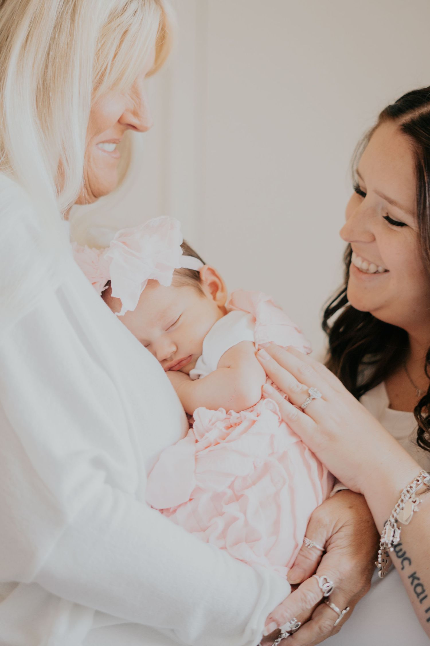 Grandmother and mother smiling while holding a sleeping newborn baby girl in pink.