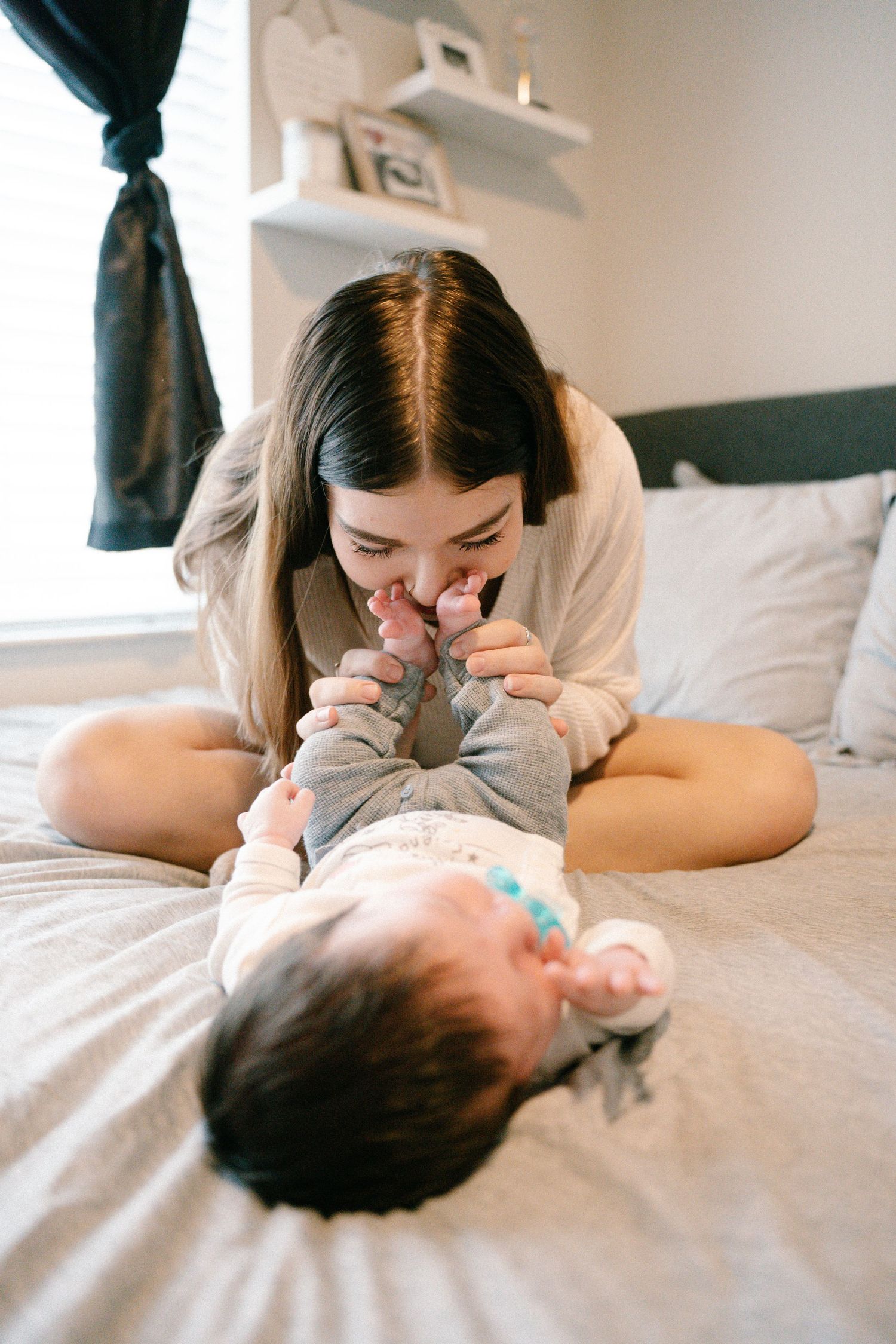 Happy mother lying on bed playfully kissing her baby's feet during bonding time.