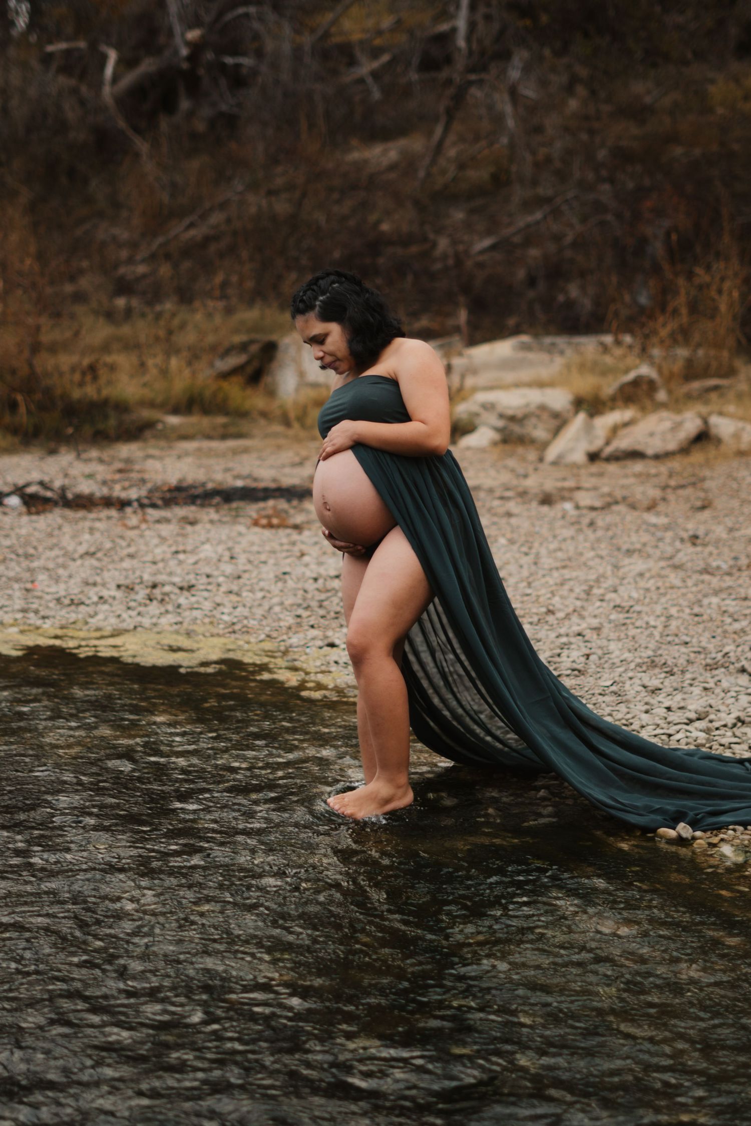 Pregnant woman standing in a shallow creek wrapped in flowing green fabric for a maternity photo session.