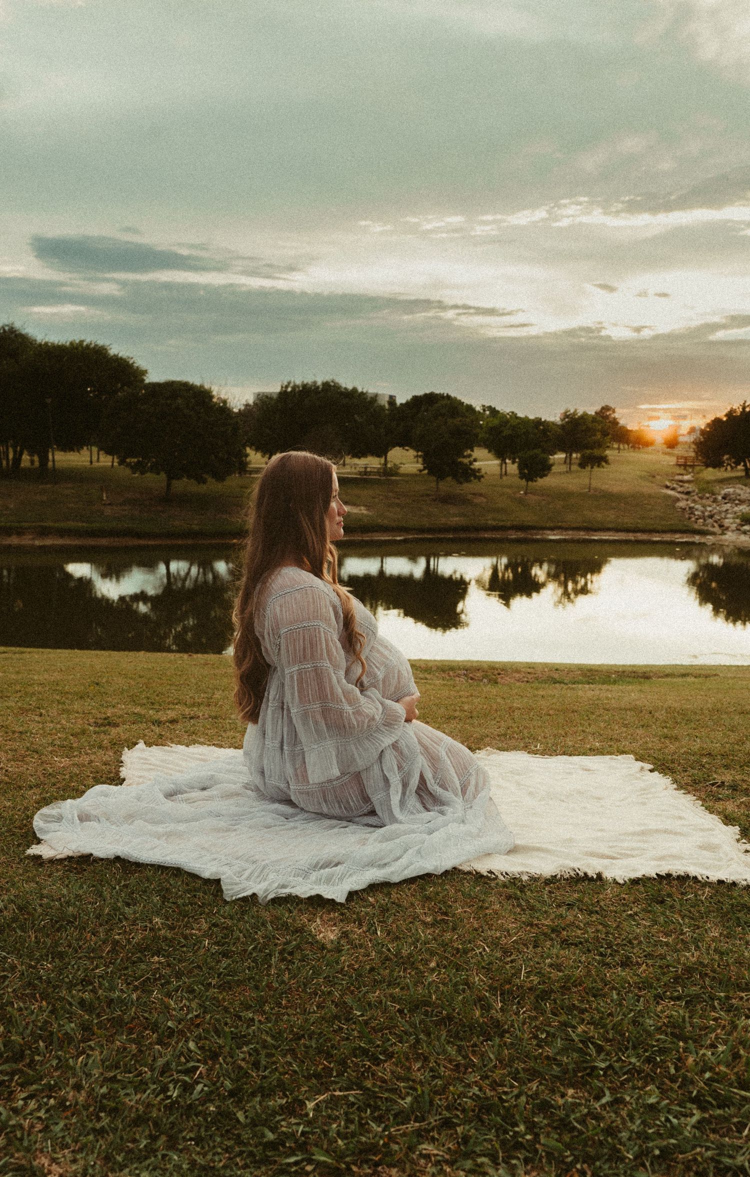 Pregnant woman in white lace dress sitting on blanket by peaceful pond at golden sunset.