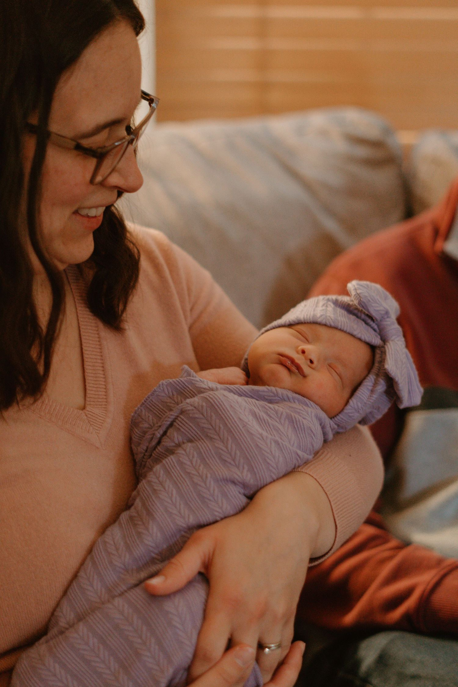 Smiling mother with glasses holding sleeping newborn in purple swaddle and bow hat.