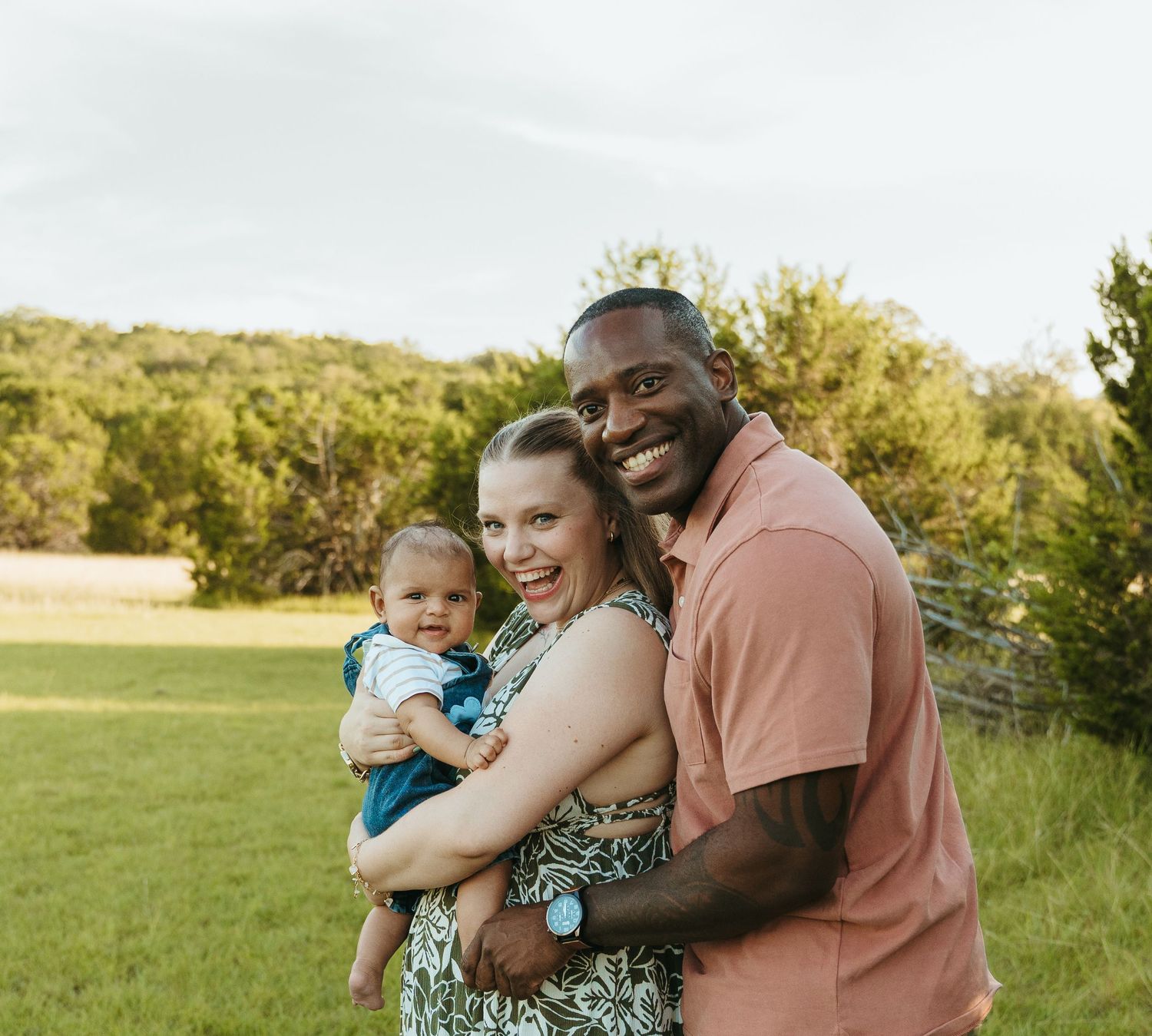 Happy interracial family of three posing outdoors in green field, father holding baby boy.