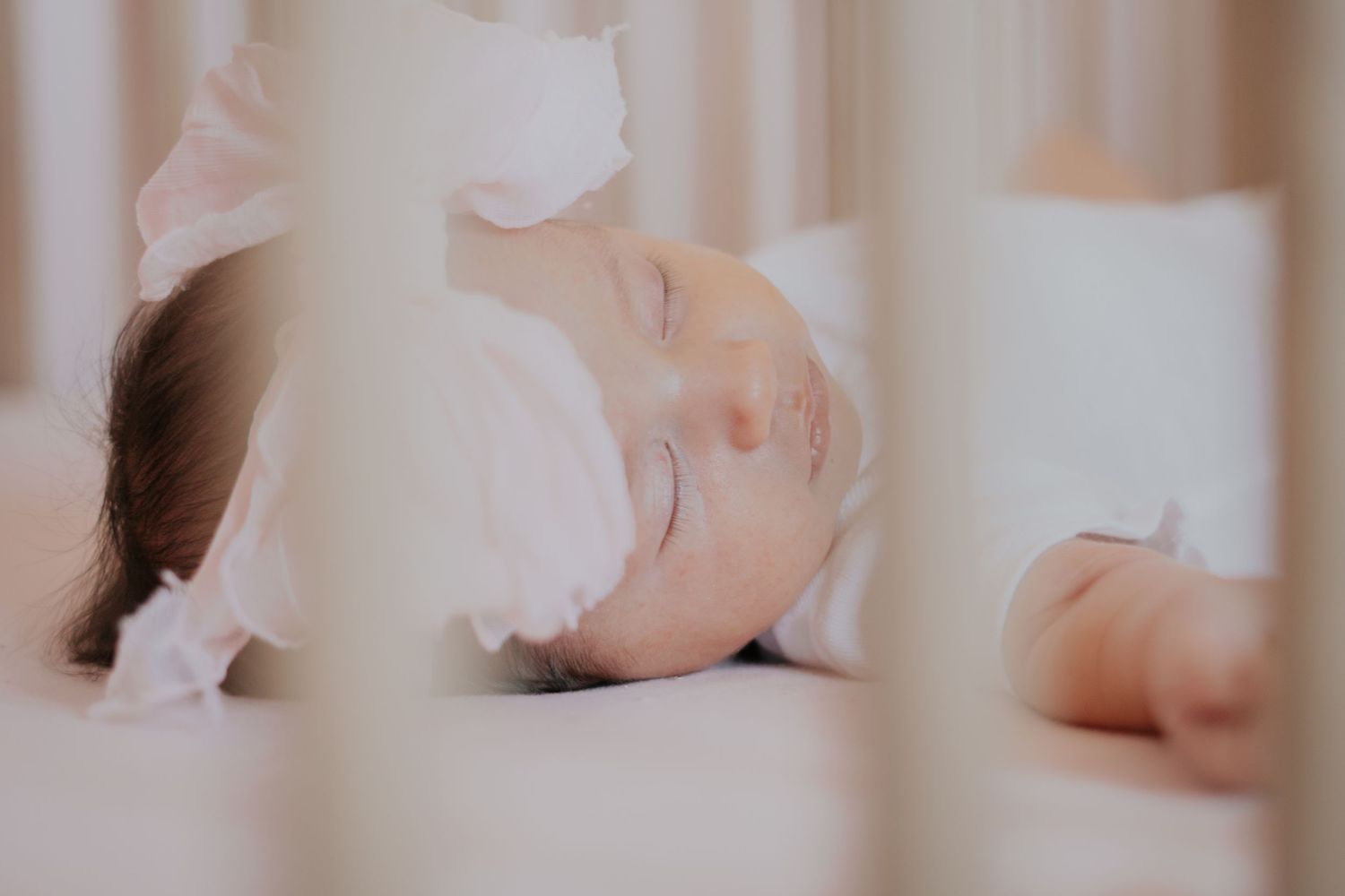 Peaceful newborn baby sleeping in a white crib wearing a white bow headband.