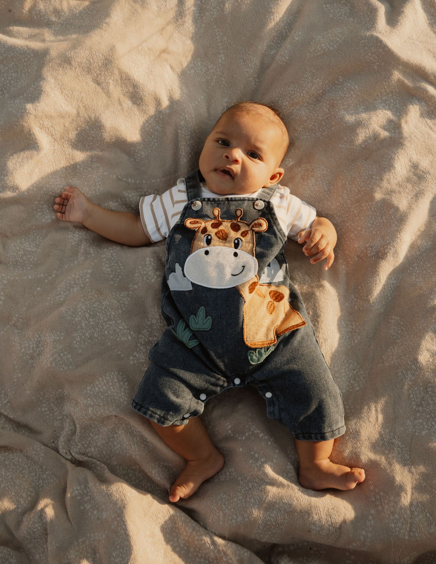 Adorable baby lying on blanket wearing giraffe overalls, looking up at camera in warm light.