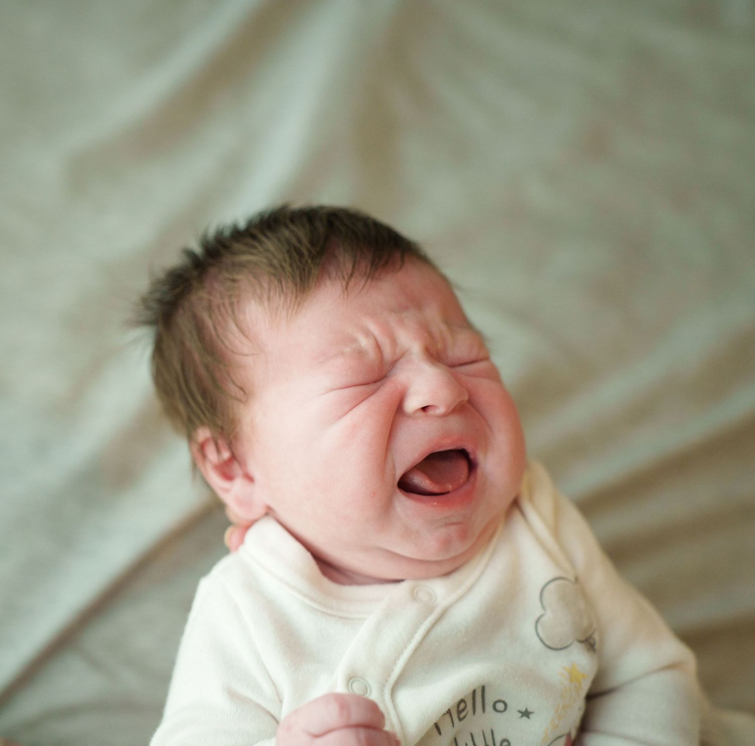 Close-up of a crying newborn baby in a white onesie against a light background.