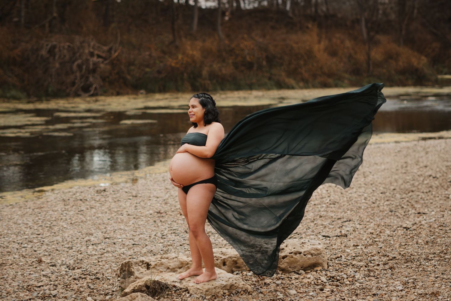 Pregnant woman in black bikini poses with flowing black fabric on rocky riverbank at dusk.