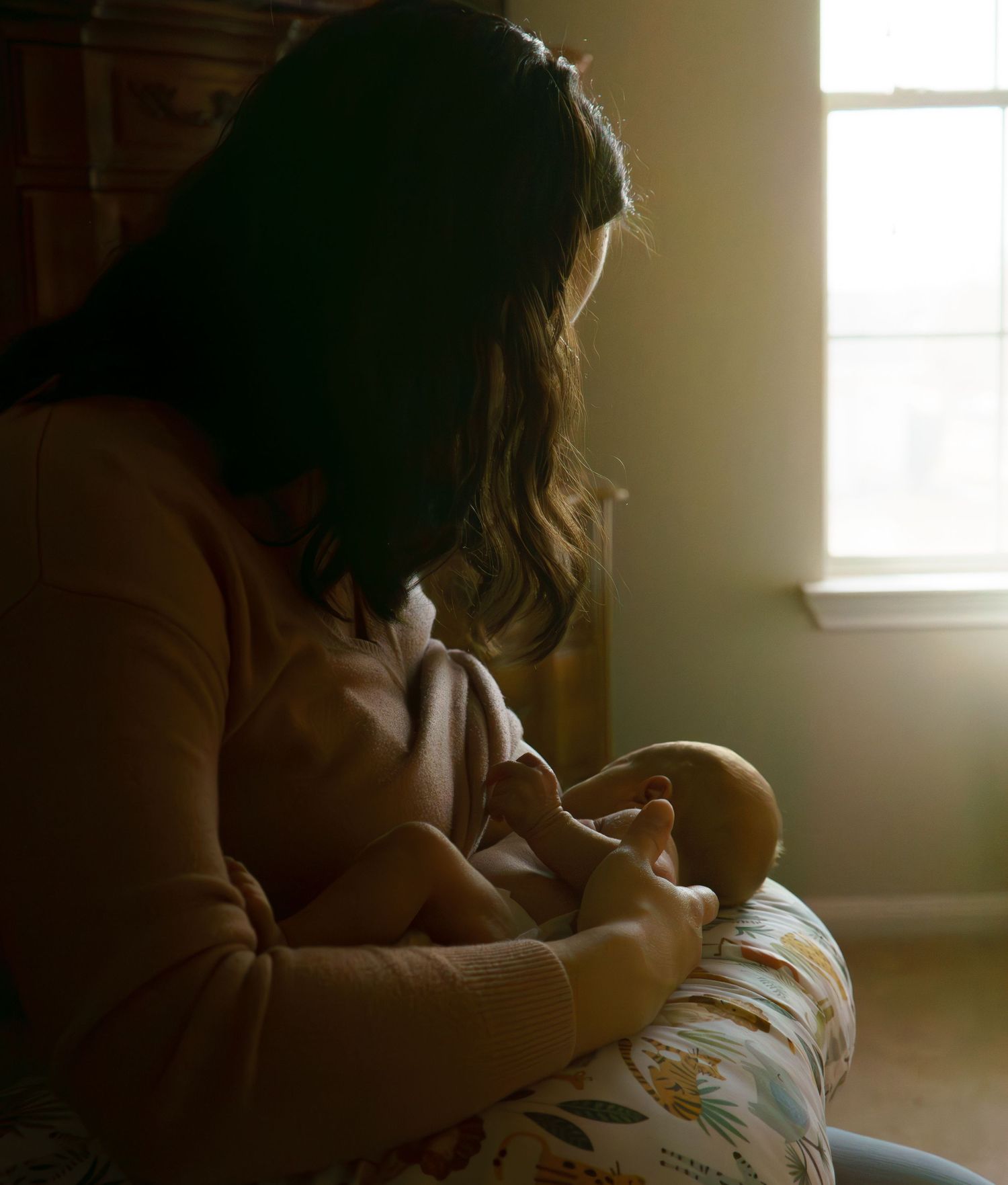Mother breastfeeding newborn baby in dimly lit room beside a bright window.