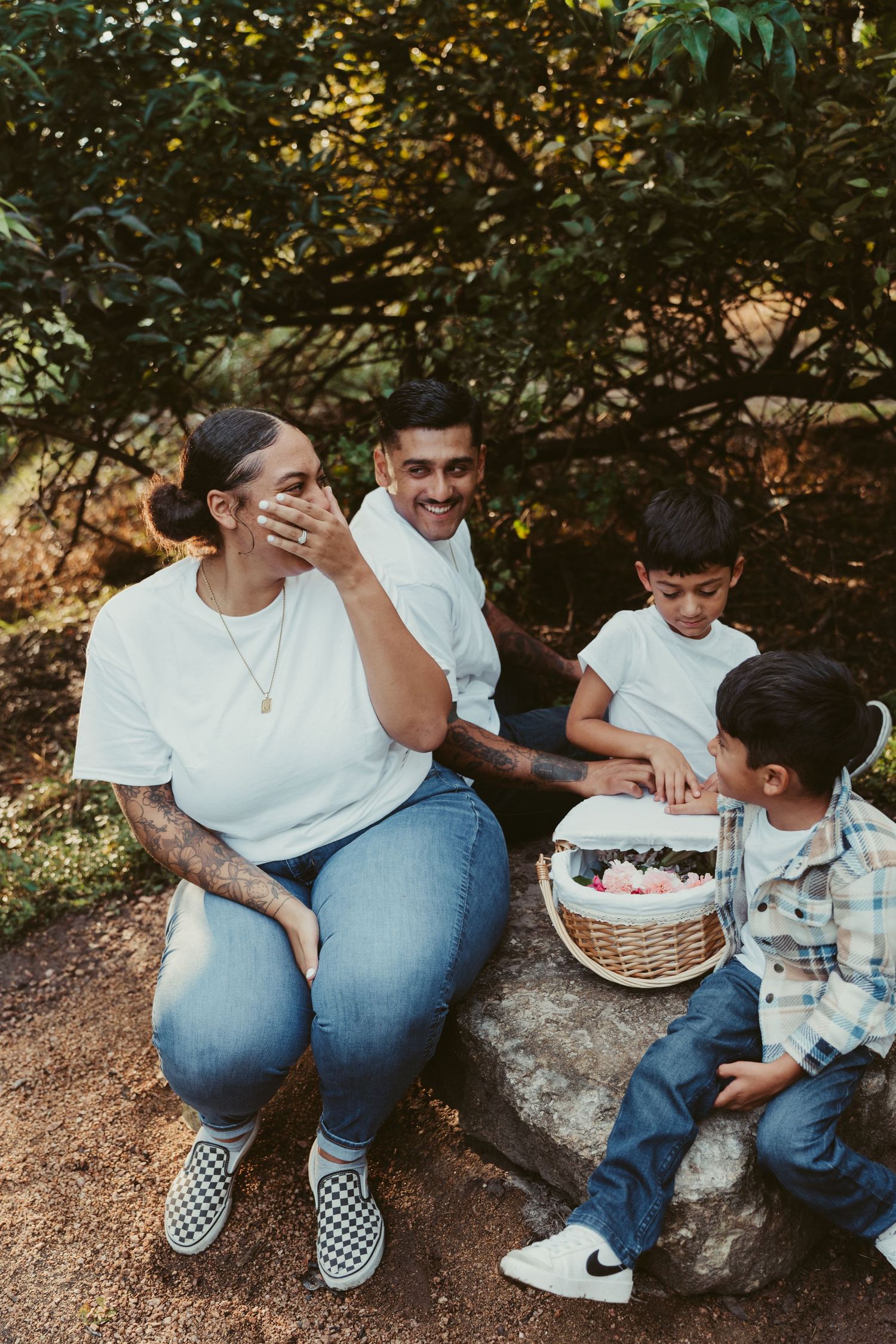 A joyful family of four sits together on a rock laughing with a flower basket in nature.