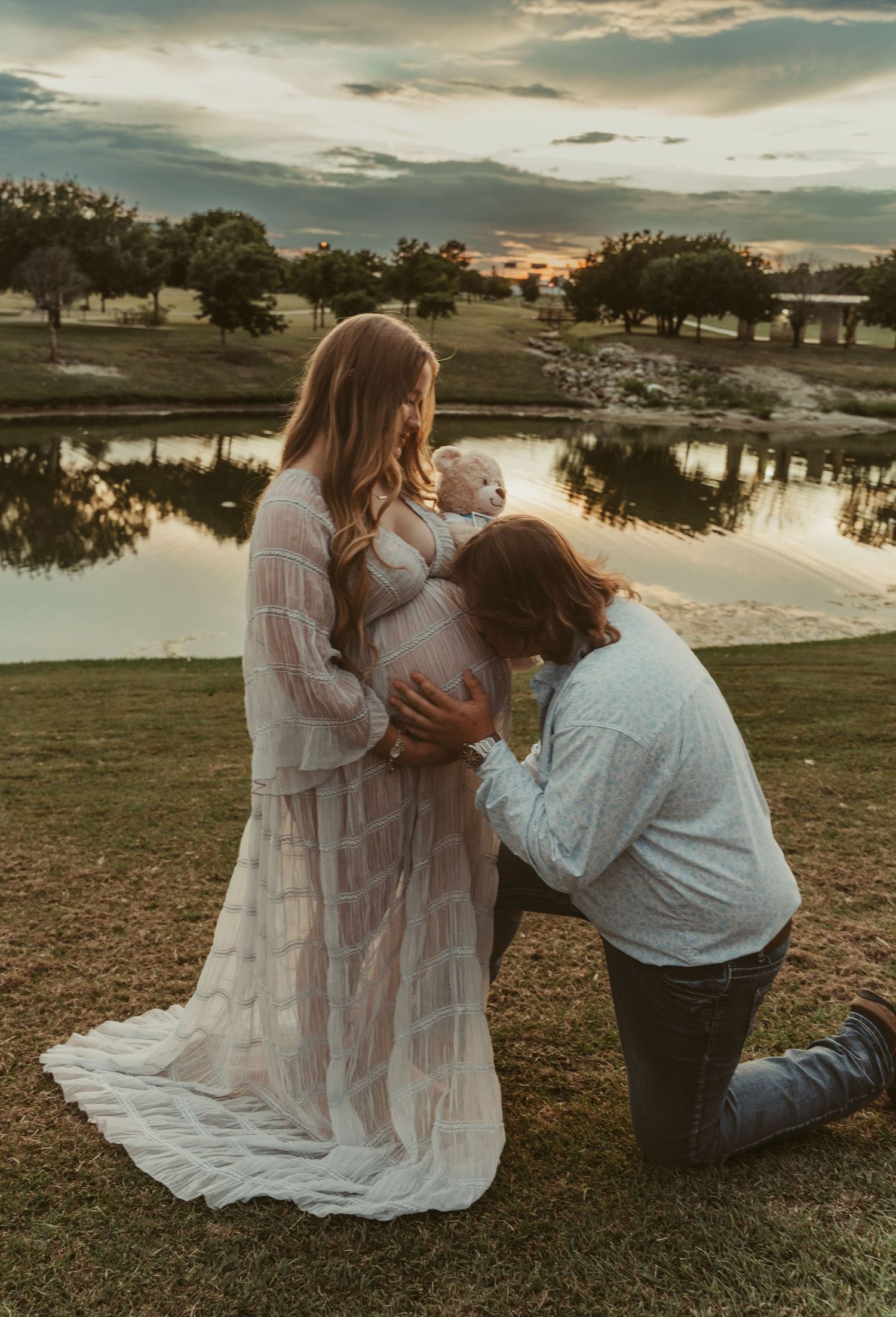 Couple sharing a tender maternity moment at sunset by a reflective pond, man kneeling kissing baby bump.