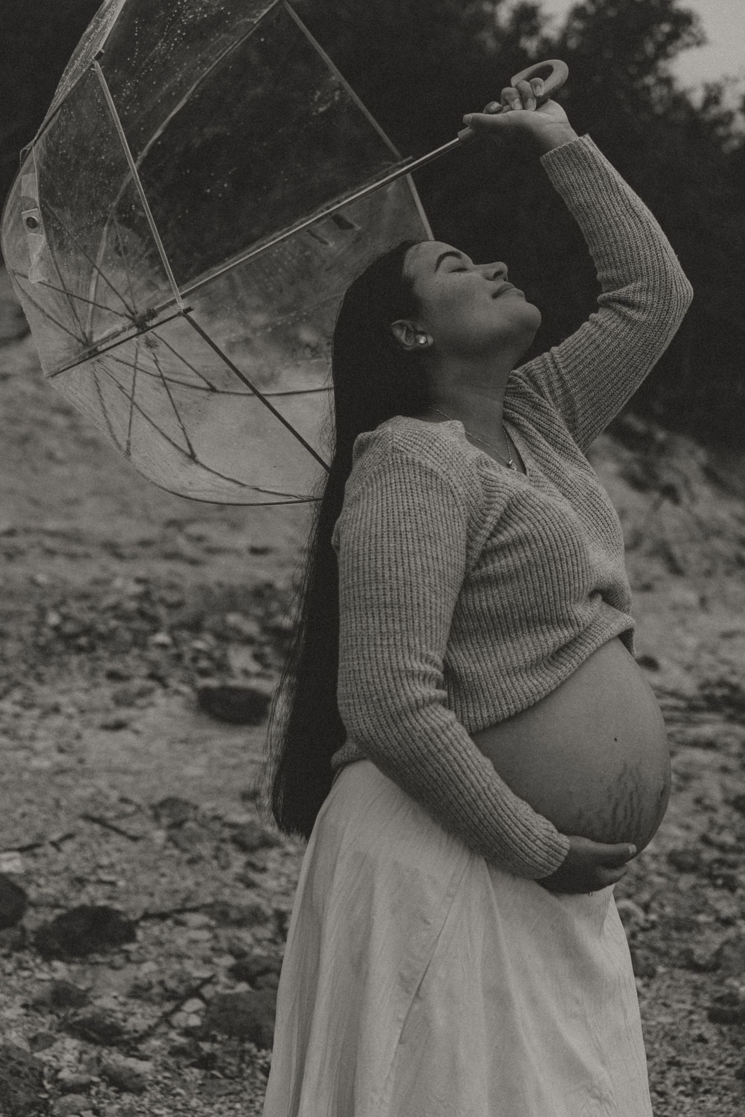 Black and white photo of pregnant woman raising clear umbrella outdoors, cradling baby bump.