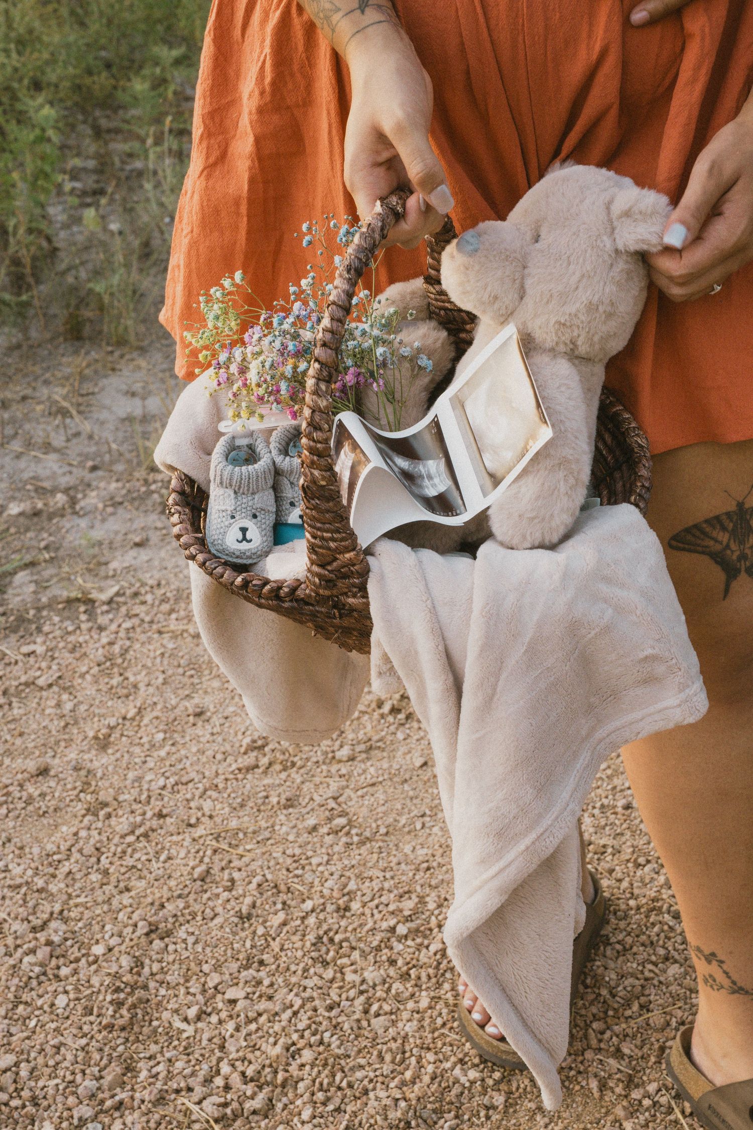 Woman in orange dress holding a wicker basket with baby shoes, toy, and blanket.