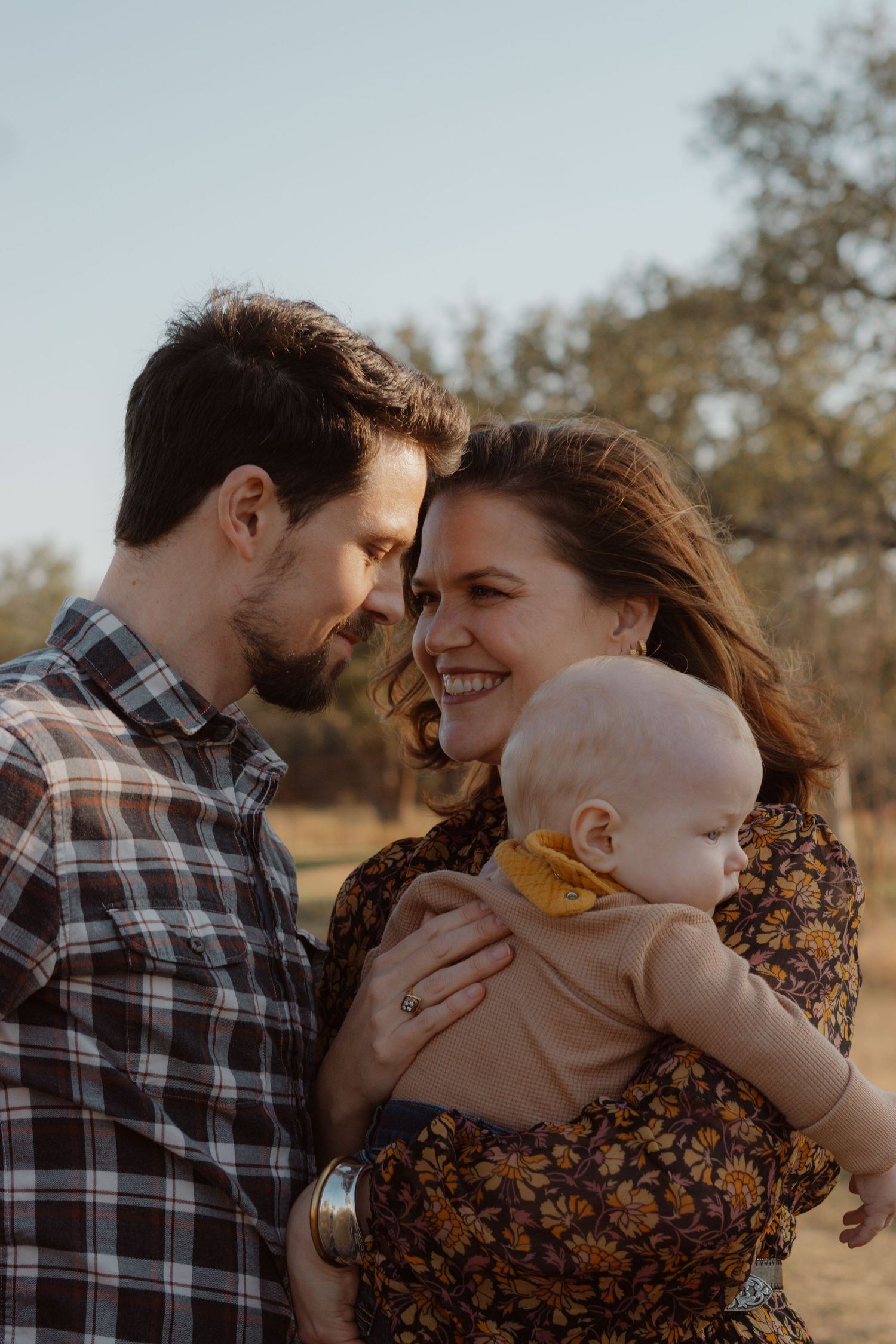 Smiling couple shares tender moment while holding baby during golden hour outdoor family portrait.