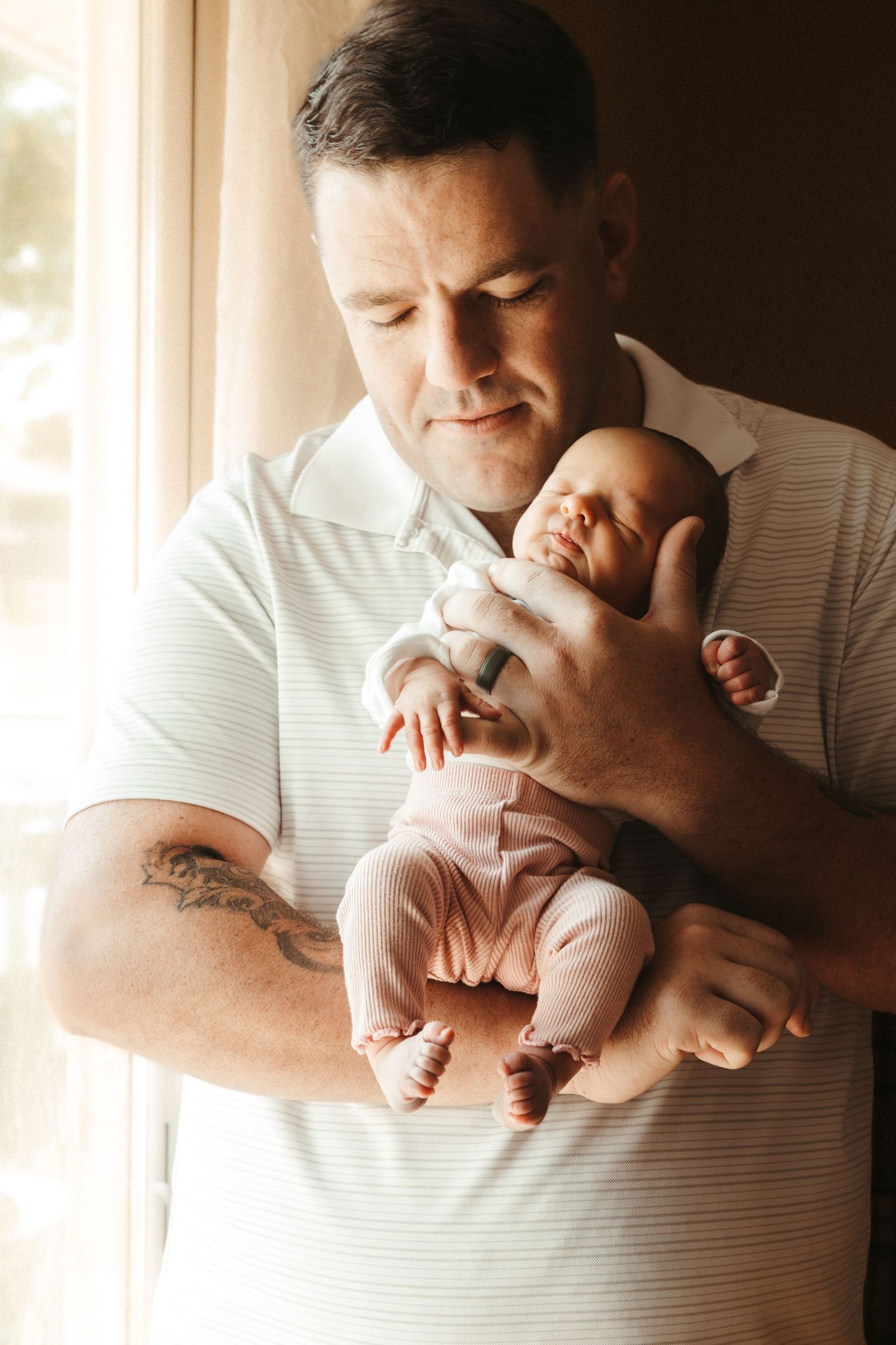 A tattooed father tenderly holds his sleeping newborn baby near a sunlit window.