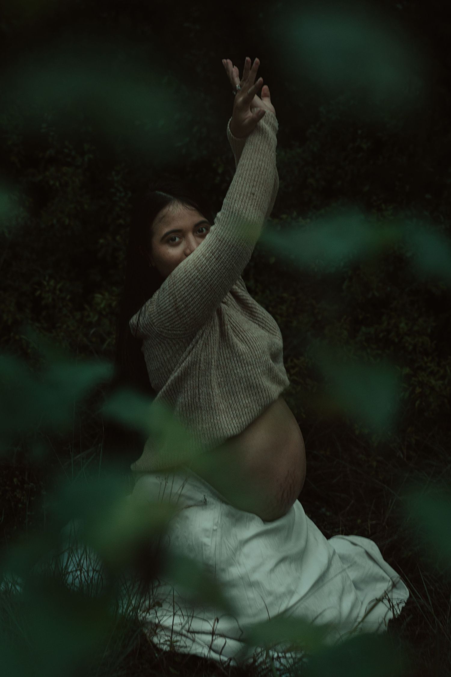 Pregnant woman reaching upward with one arm while seated among green foliage in forest.