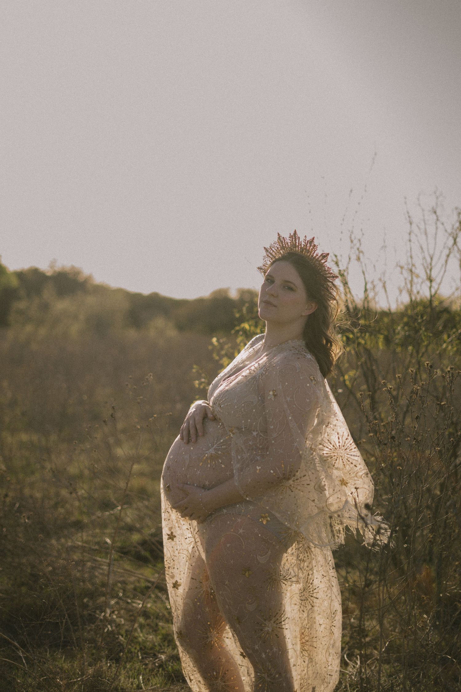 Pregnant woman wearing lace wrap and crown poses in sunlit green field during golden hour.