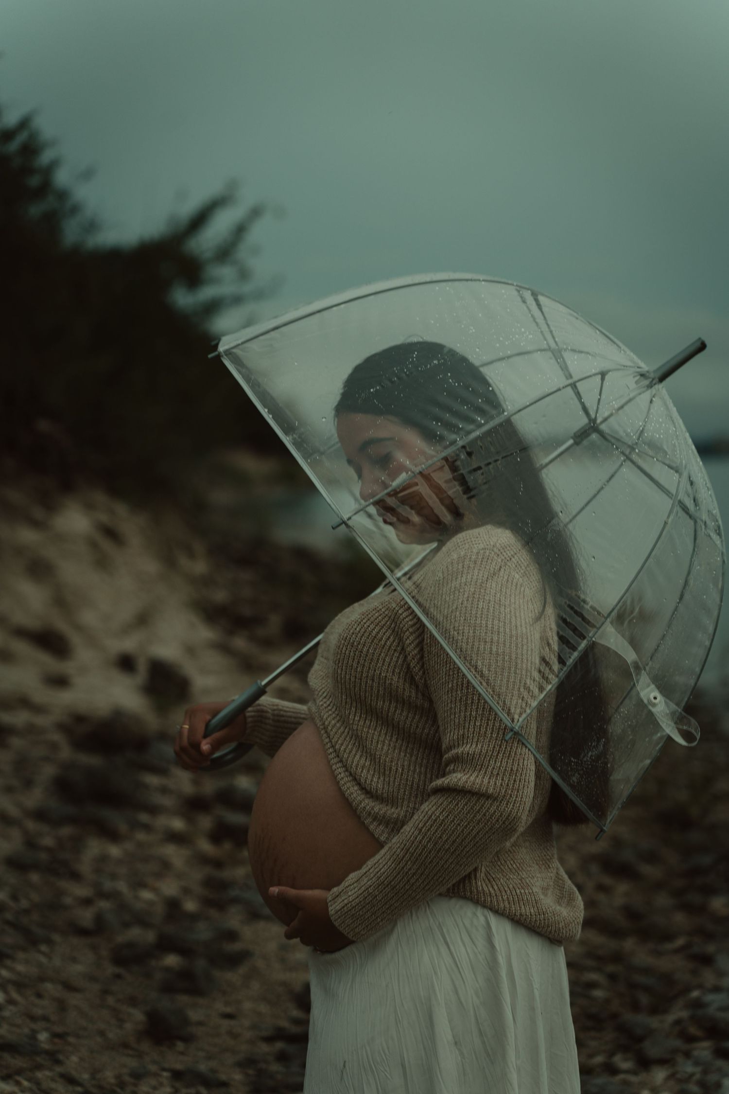 Pregnant woman in beige sweater holding clear umbrella outdoors on rocky terrain in moody weather.