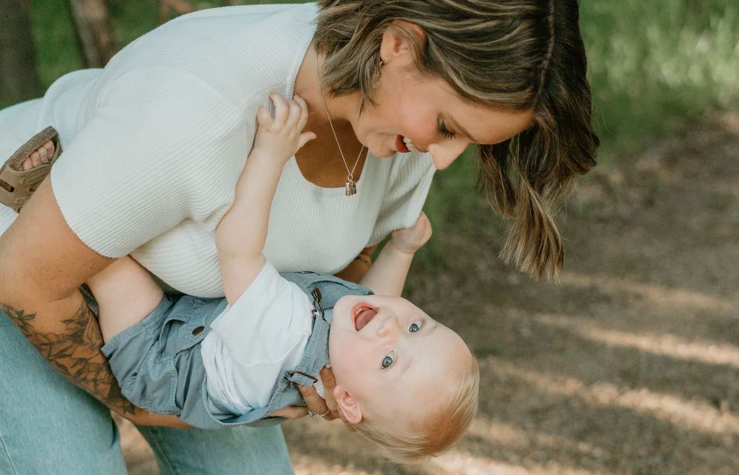 Joyful mother leaning over laughing baby outdoors in wooded setting.