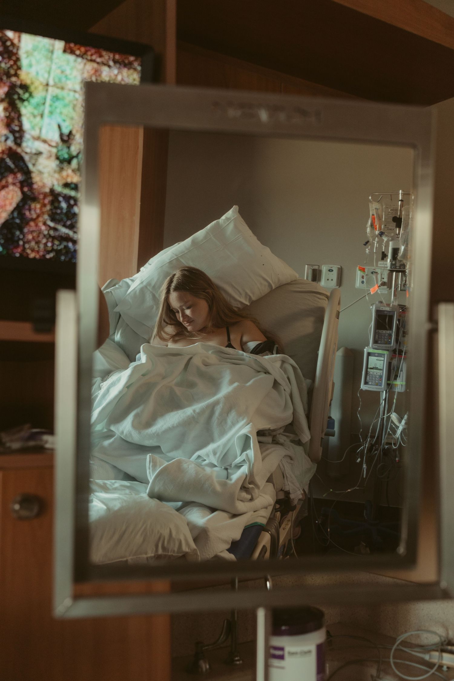 A new mother rests in a hospital bed holding her newborn, reflected in a mirror.