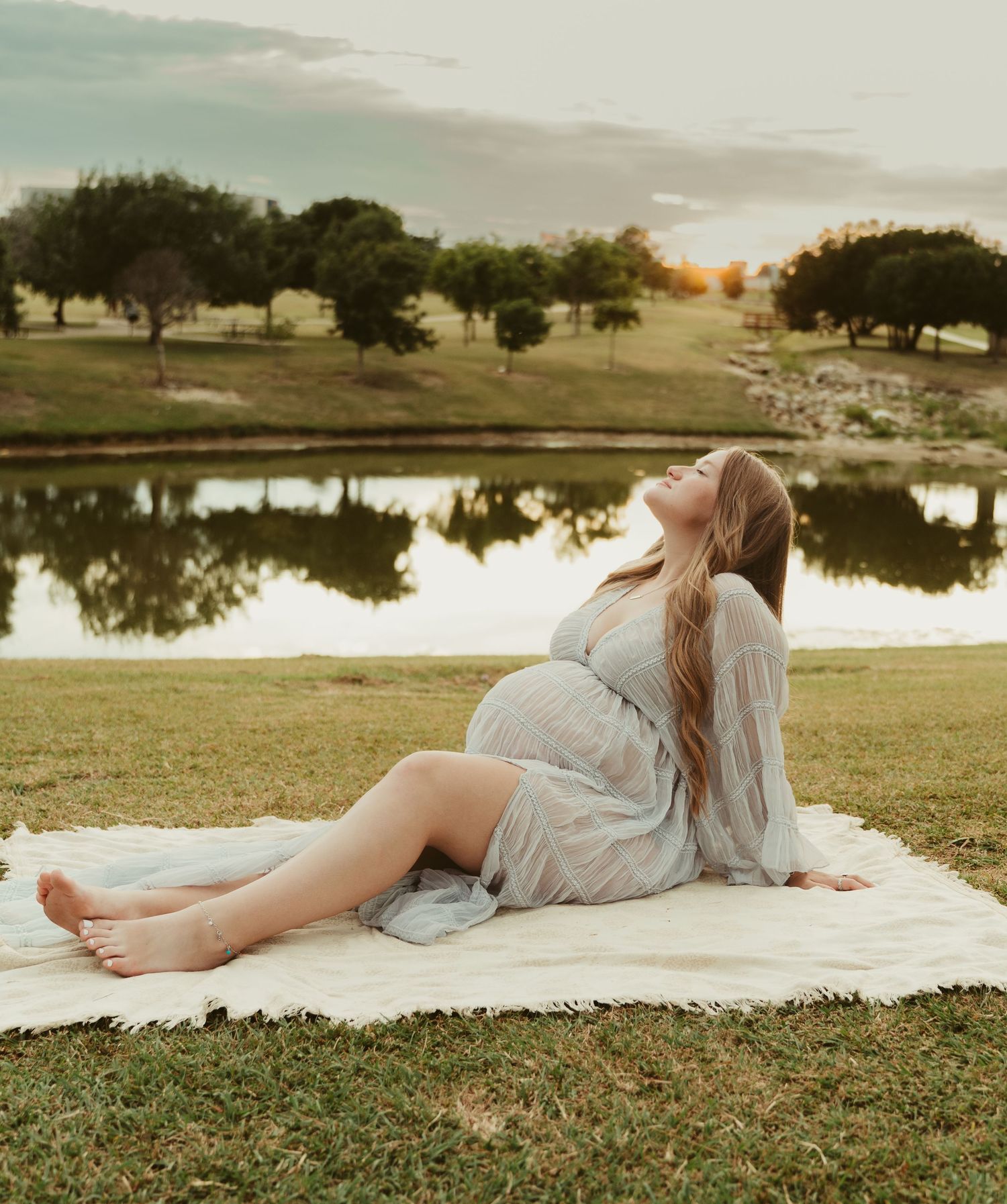 Barefoot pregnant woman in sheer dress sitting on blanket by lake, tilting head back at sunset.