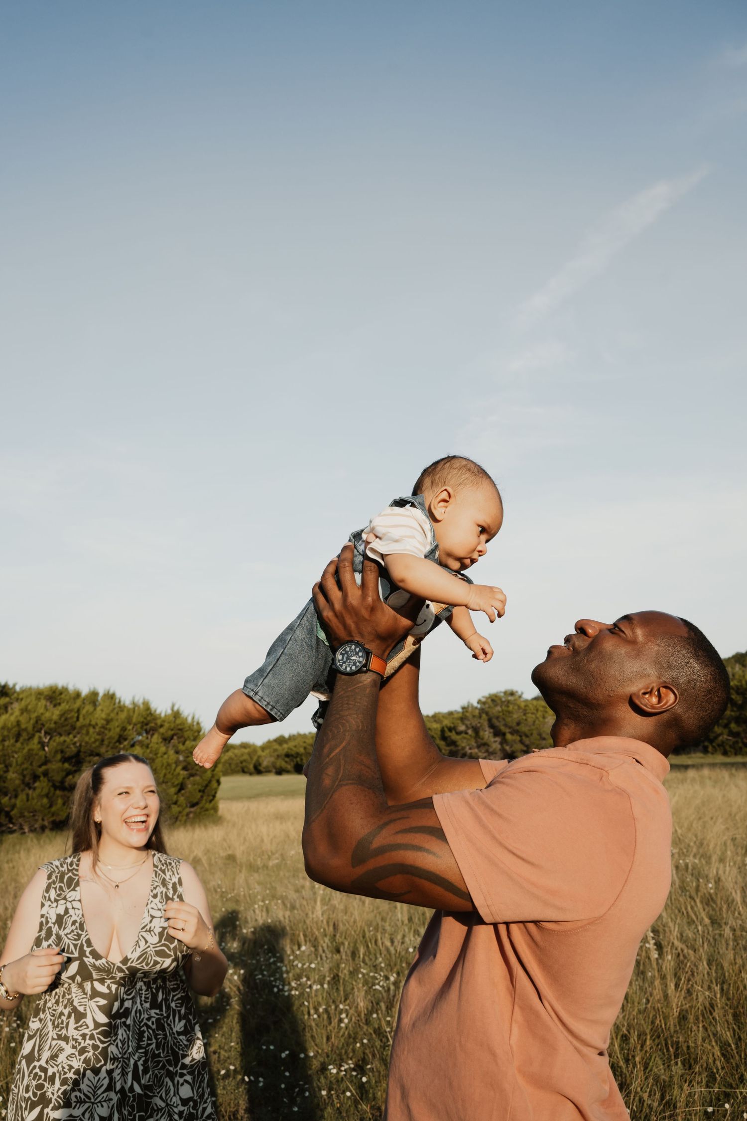 Father joyfully lifting baby into the air while mother laughs in a sunny green field.