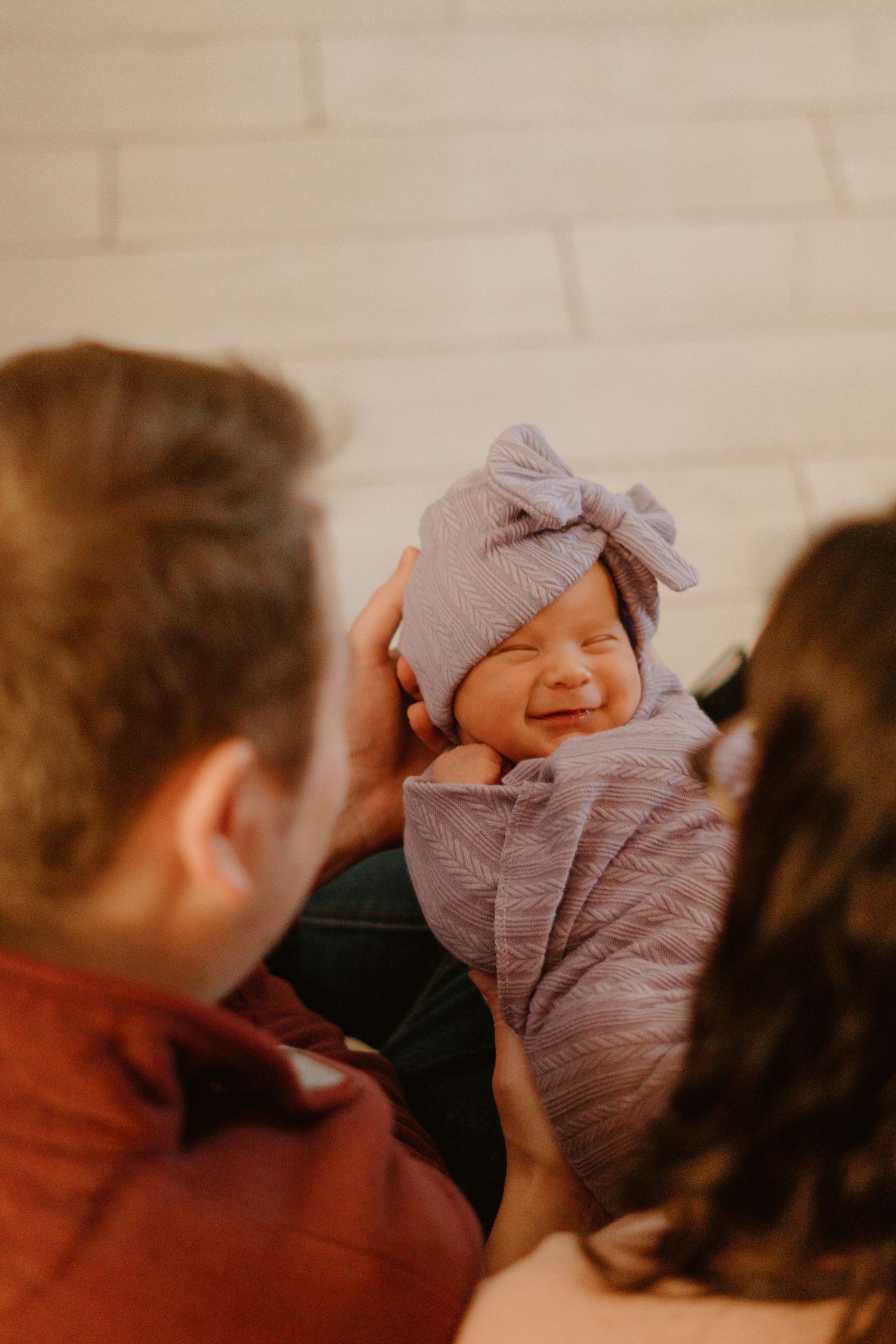 Smiling newborn baby in purple swaddle and bow hat held by parents.
