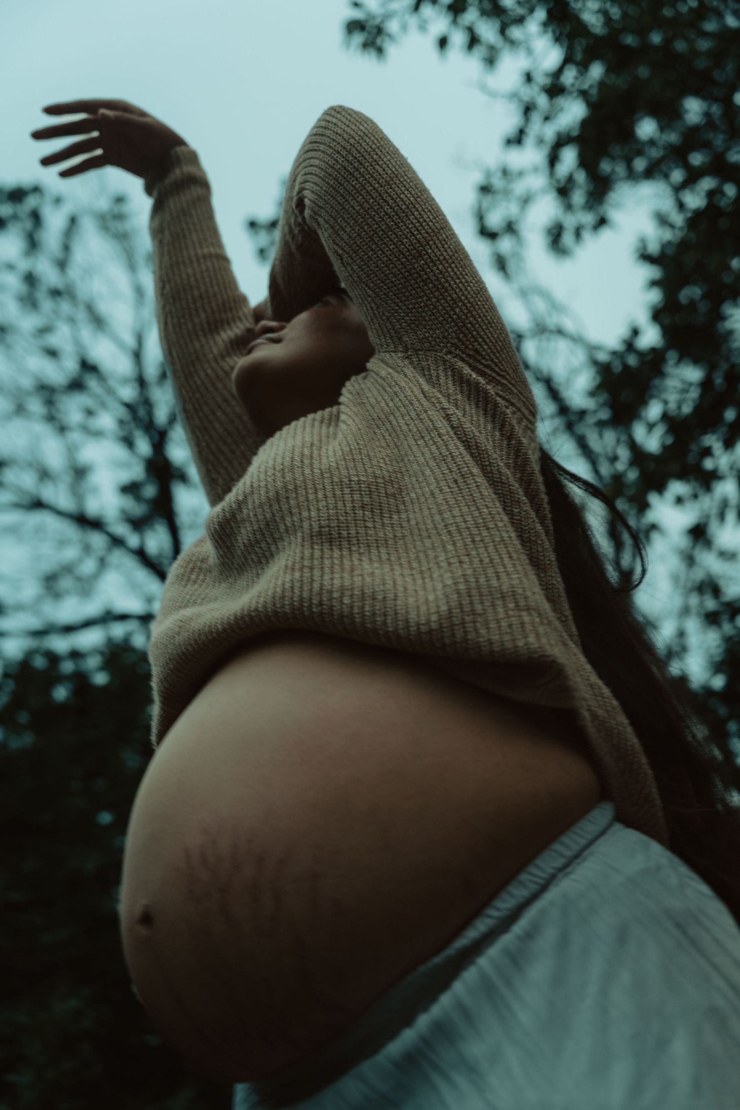 Low angle view of pregnant woman in beige sweater raising arm toward sky among trees.